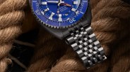 Black metal wristwatch with a blue diver GMT dial and a linked bracelet resting on thick brown rope.