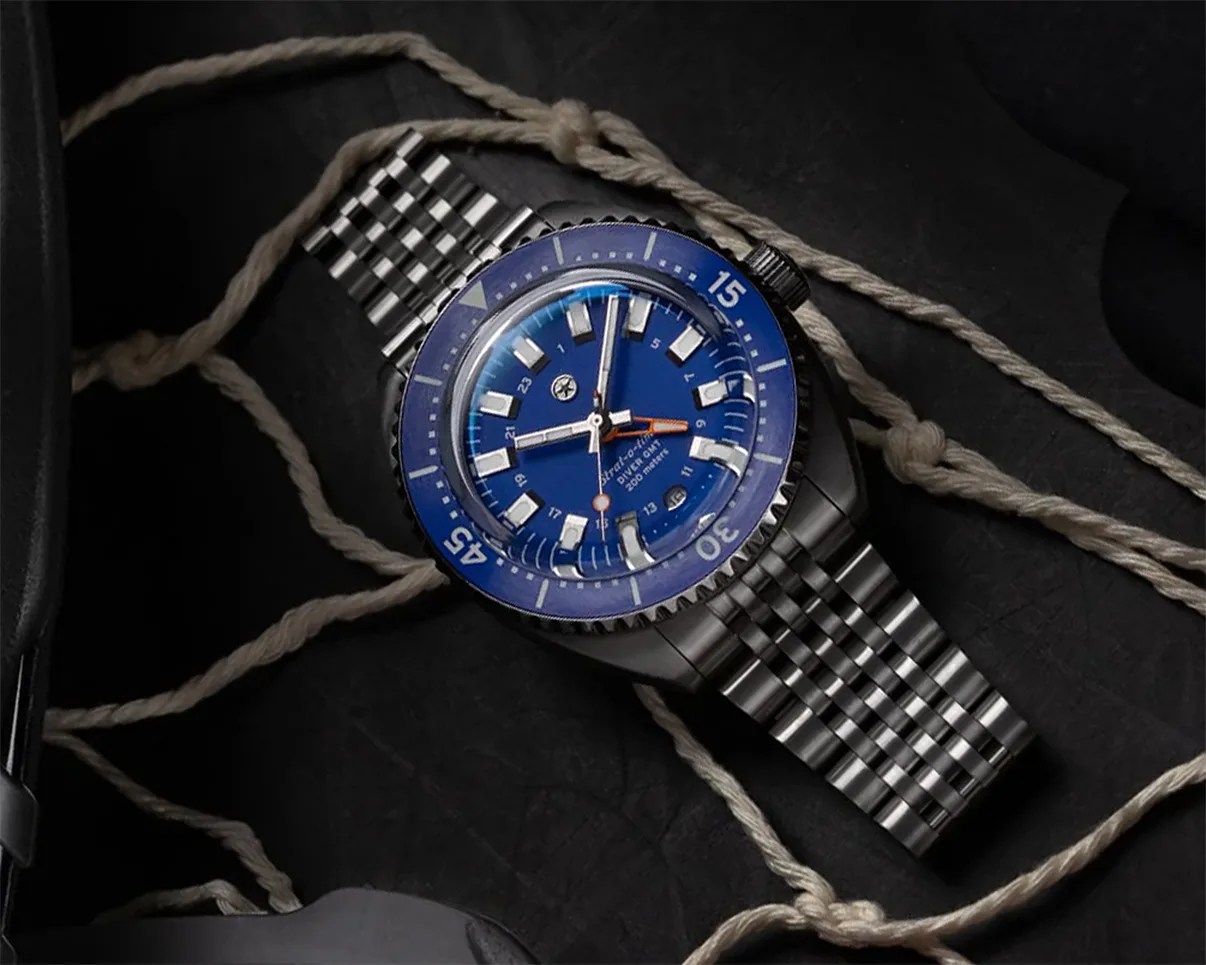 Stainless steel dive watch with blue bezel and dial, luminous markers, and metal bracelet on a dark surface with rope.