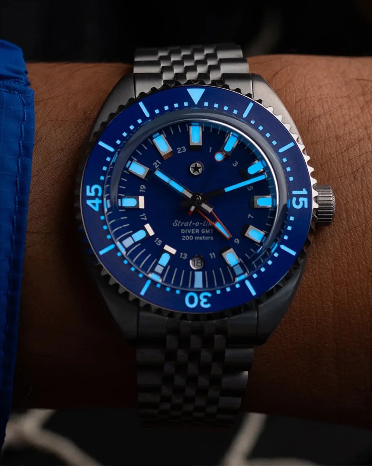 Blue dial diver GMT watch with luminous markers and a stainless steel bracelet on a wrist.