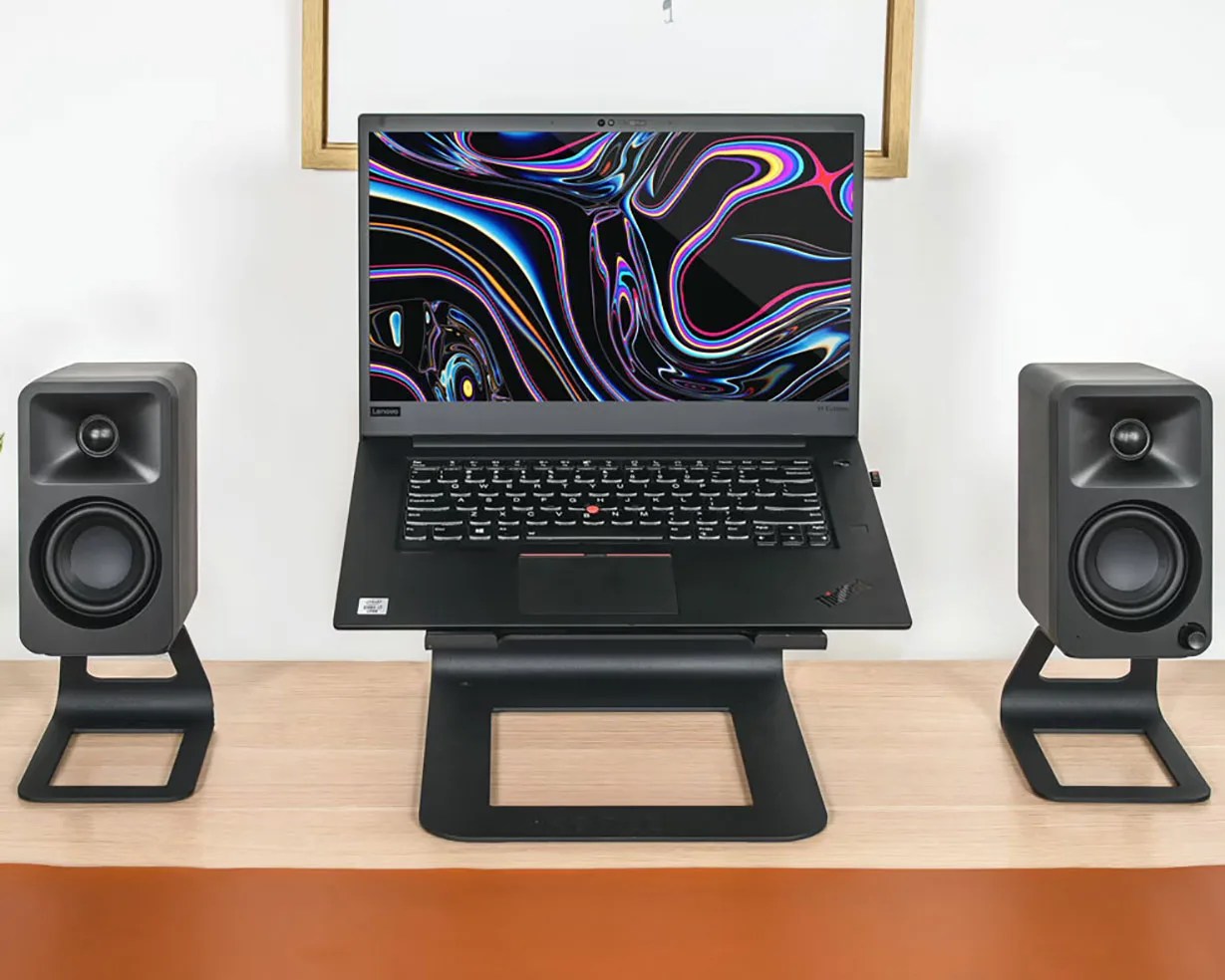 Black Lenovo laptop on a black stand flanked by two black desktop speakers on a wooden desk.