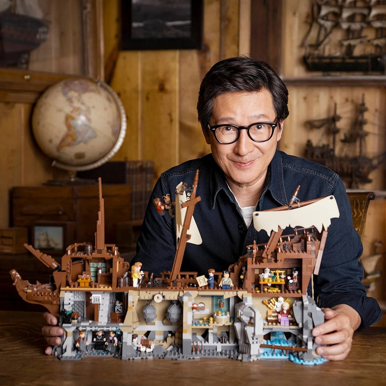 Person wearing glasses holding a detailed LEGO pirate ship model with interior scenes on a wooden table.
