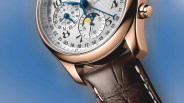 Close-up of a luxury watch with a rose gold case, white textured dial, blue hands, moon phase indicator, and brown leather strap.