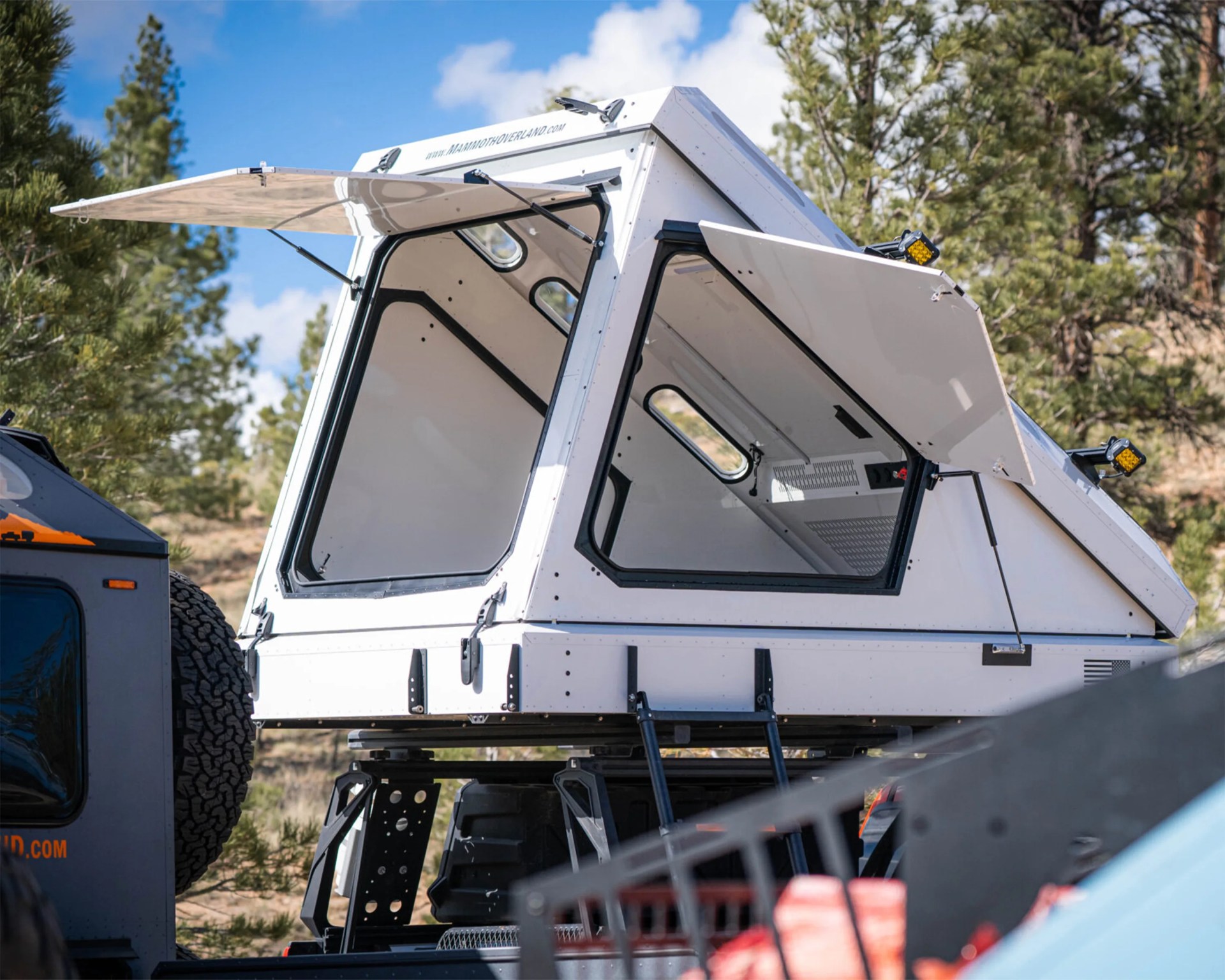 White rooftop camper shell with open side and rear windows mounted on a vehicle in a forested area.
