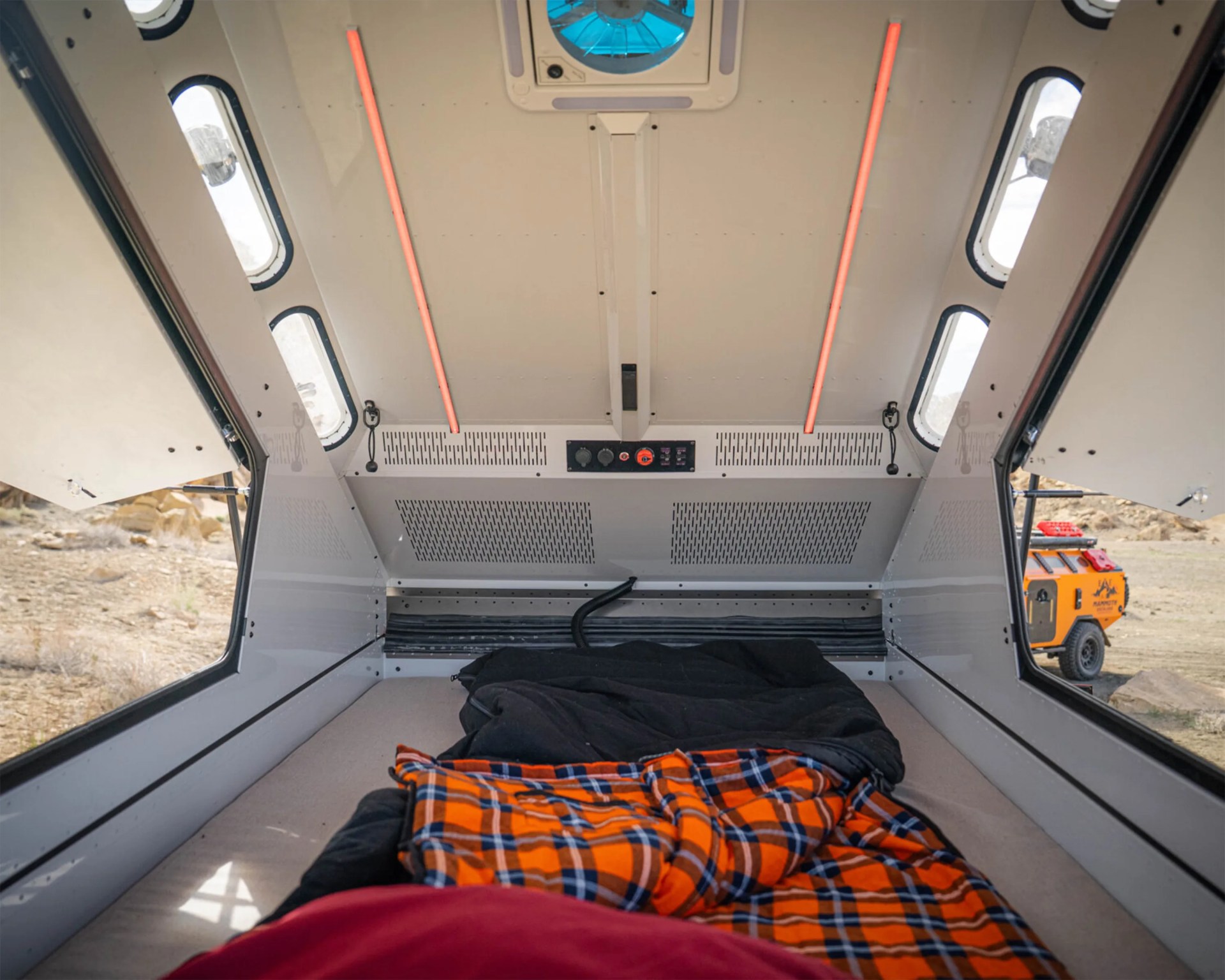 Interior view of a compact camper with a bed, orange plaid blanket, and open side windows showing a rugged outdoor setting.