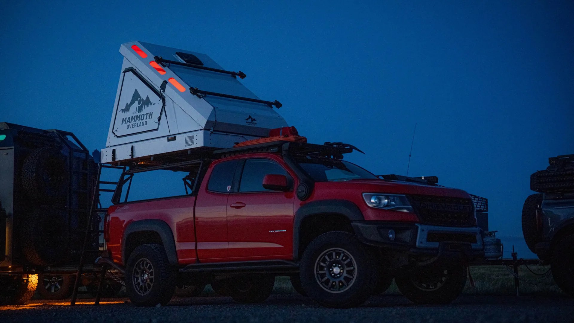 Red Chevrolet pickup truck with a white Mammoth Overland rooftop tent mounted on the bed, parked at dusk.