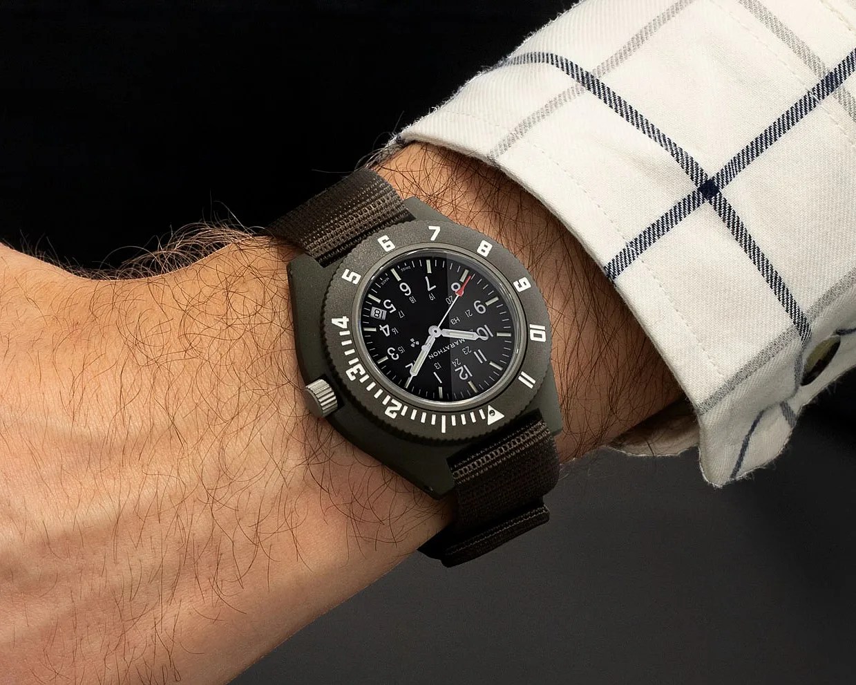 Military-style wristwatch with black dial, white numerals, and olive green case on a brown fabric strap worn on a wrist.