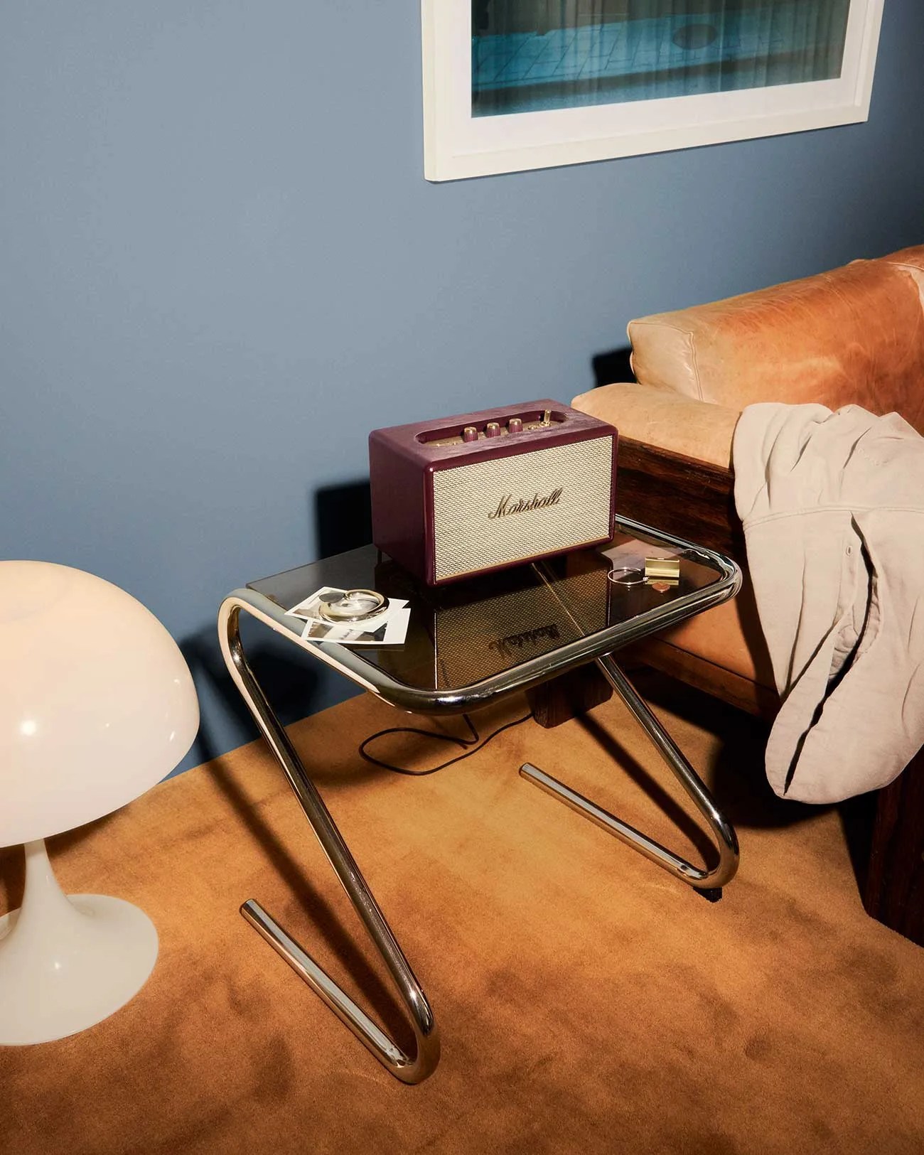 Small maroon Marshall speaker on a glass-top side table with chrome legs next to a tan leather sofa.