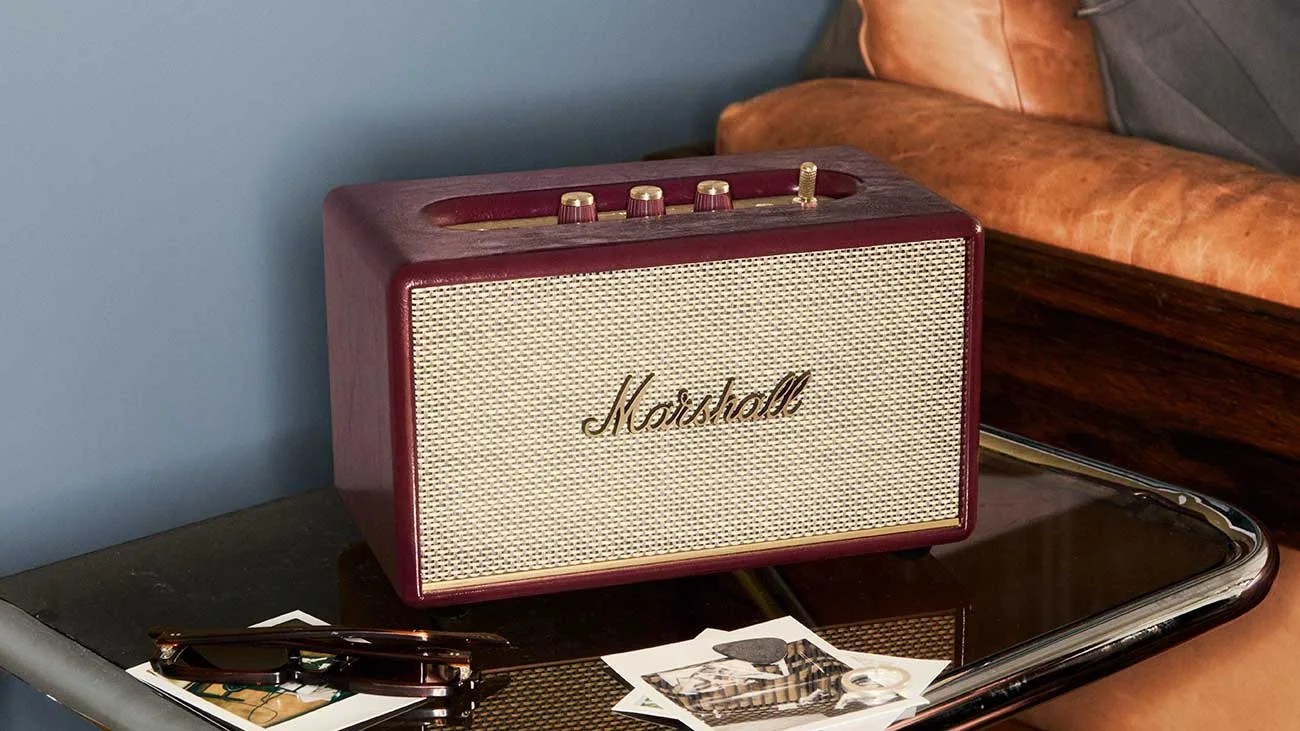 Burgundy Marshall speaker with gold knobs on a glass table next to sunglasses and photos.