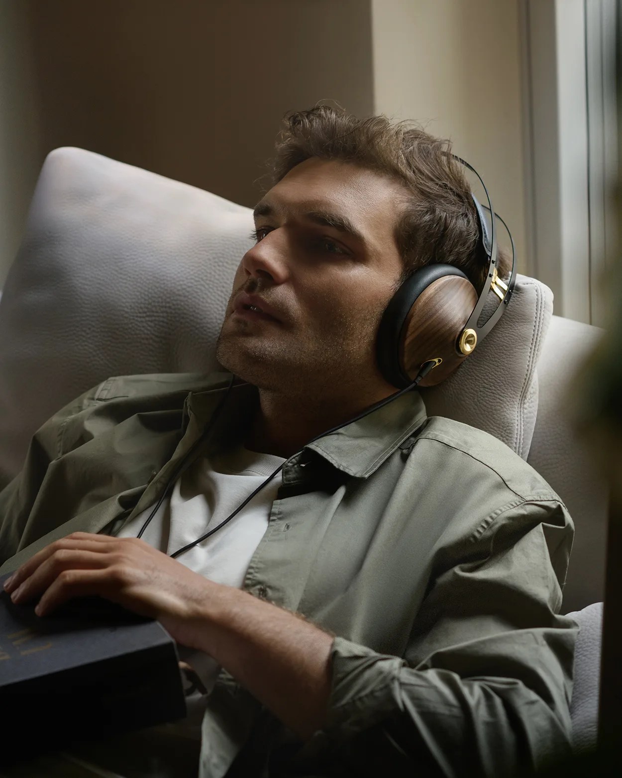Man reclining on a light gray couch wearing wooden over-ear headphones and holding a black book.
