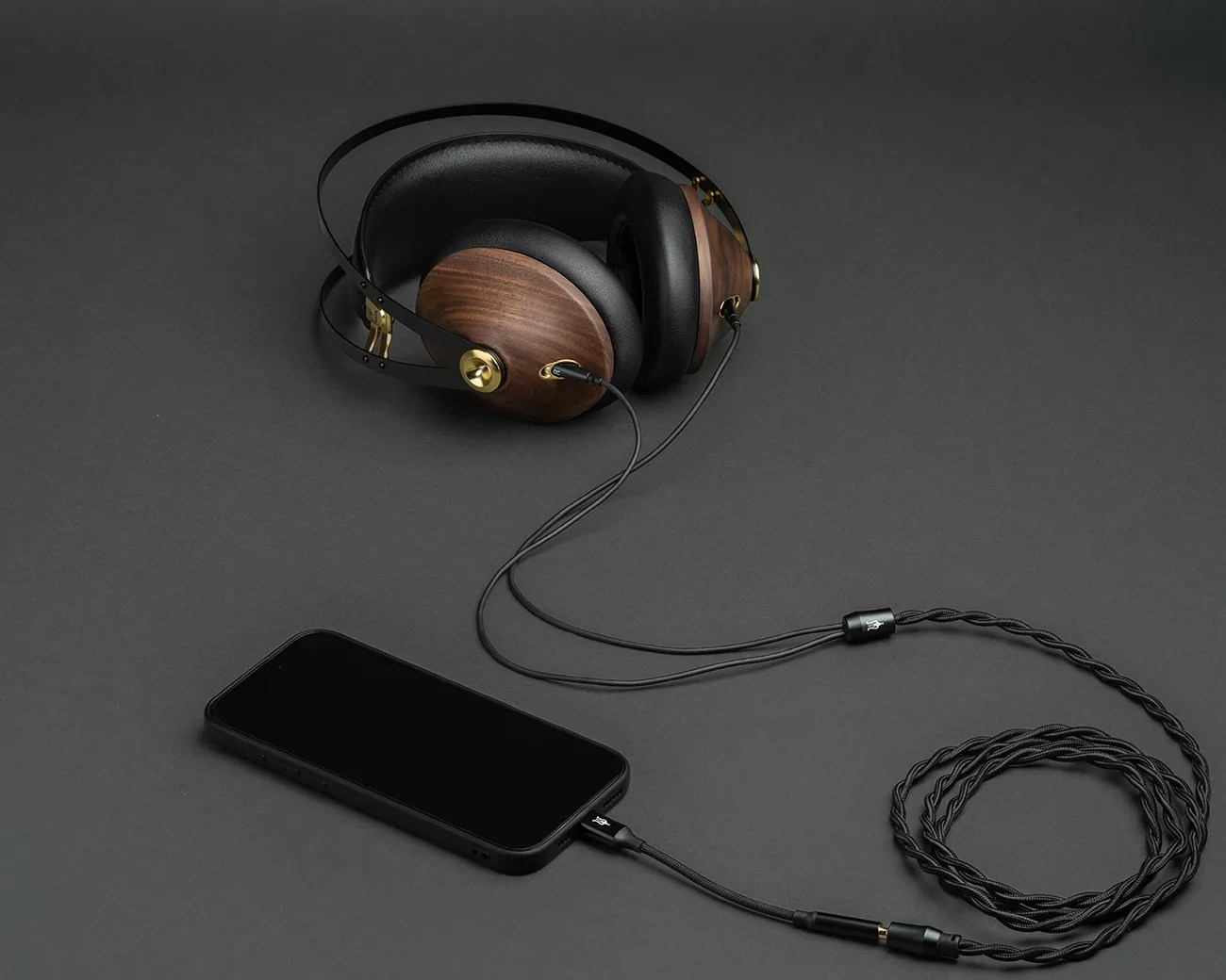 Over-ear headphones with wooden ear cups and black leather padding connected to a black smartphone by a braided cable on a dark surface.
