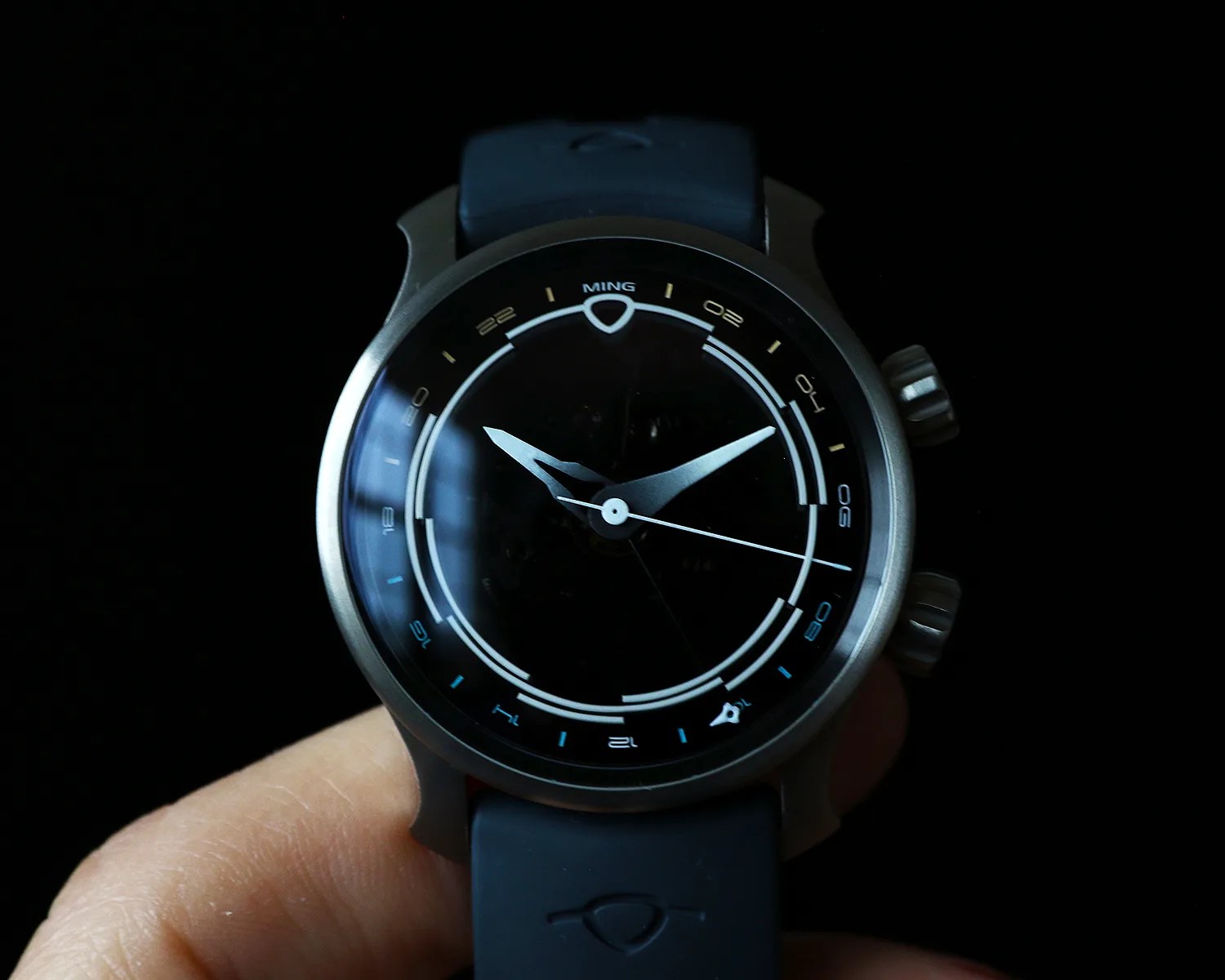 Round silver wristwatch with black dial, white hour and minute hands, and blue rubber strap held in a hand.