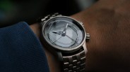 Silver metal wristwatch with a transparent dial showing internal gears, worn on a wrist with a blue sleeve.