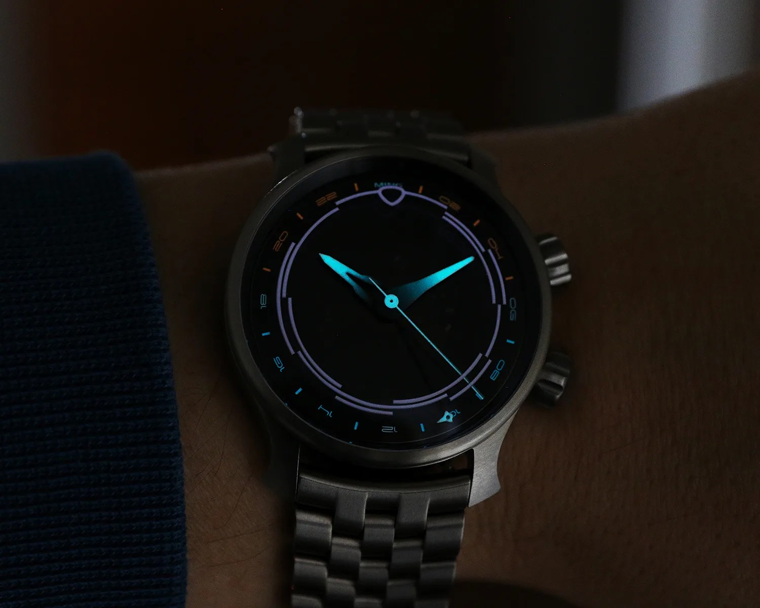 Round wristwatch with glowing blue hands and markers on a dark face, worn on a wrist.