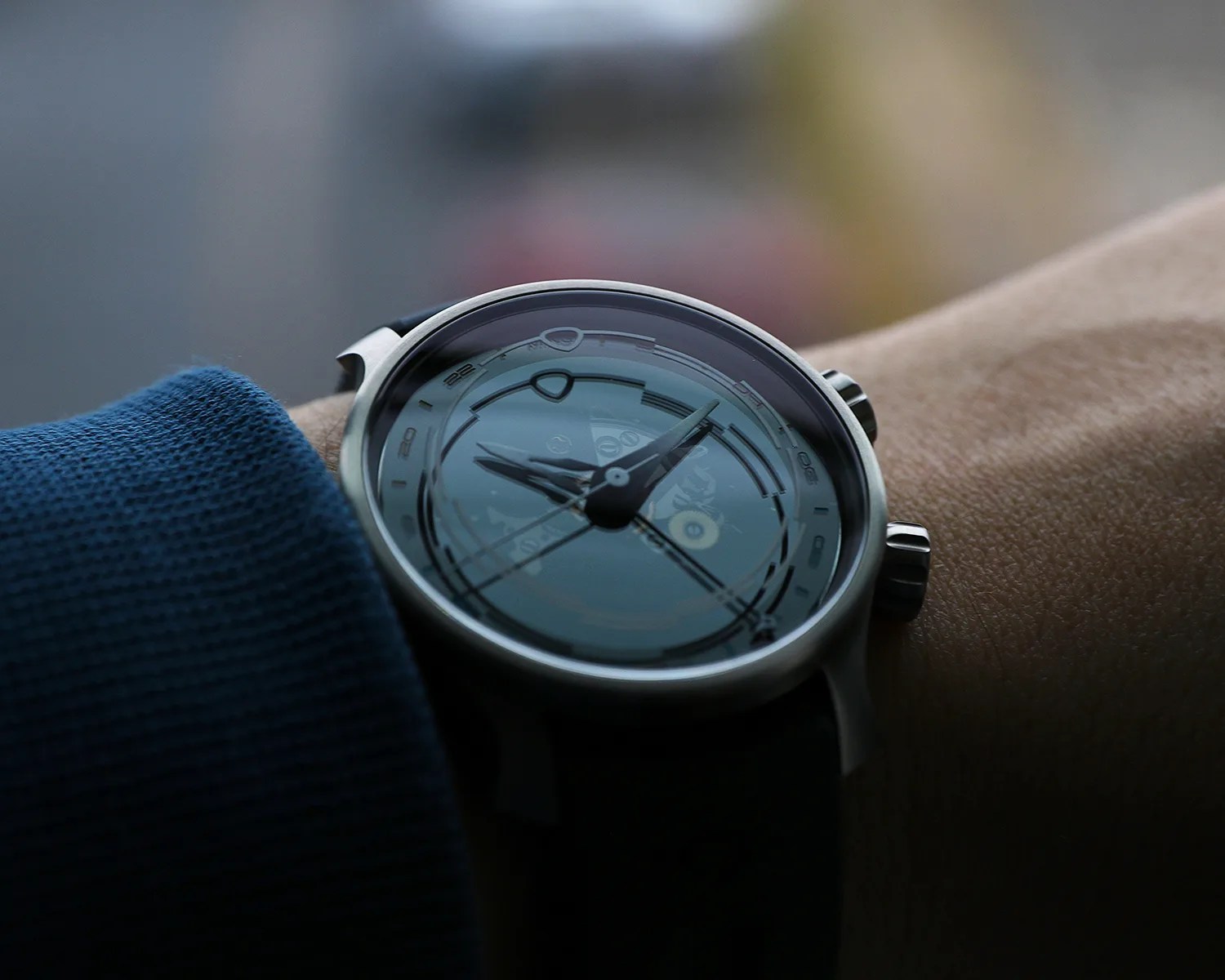 Close-up of a wristwatch with a silver case and strap worn on a wrist with a blue sleeve.