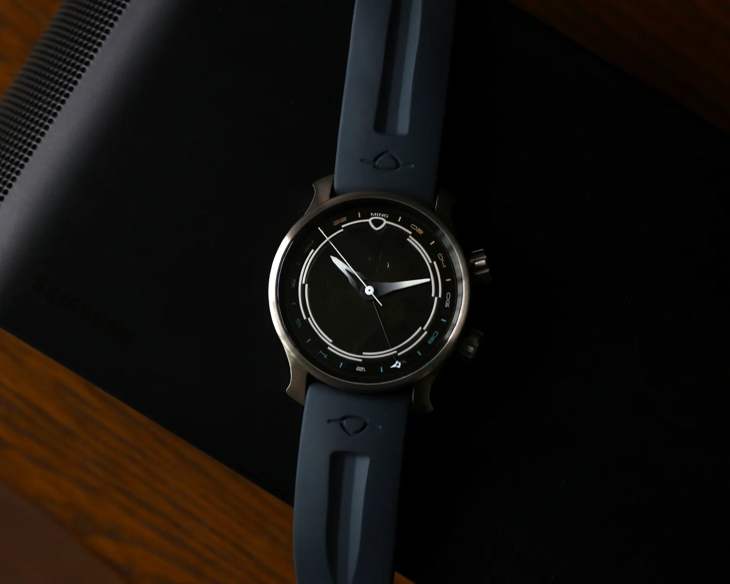 Round wristwatch with a black dial, silver case, and blue rubber strap placed on a black Samsung device.
