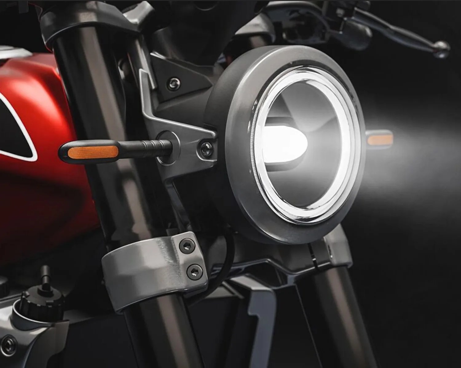 Close-up of a round motorcycle headlight with a bright beam and orange turn signals on black front forks.