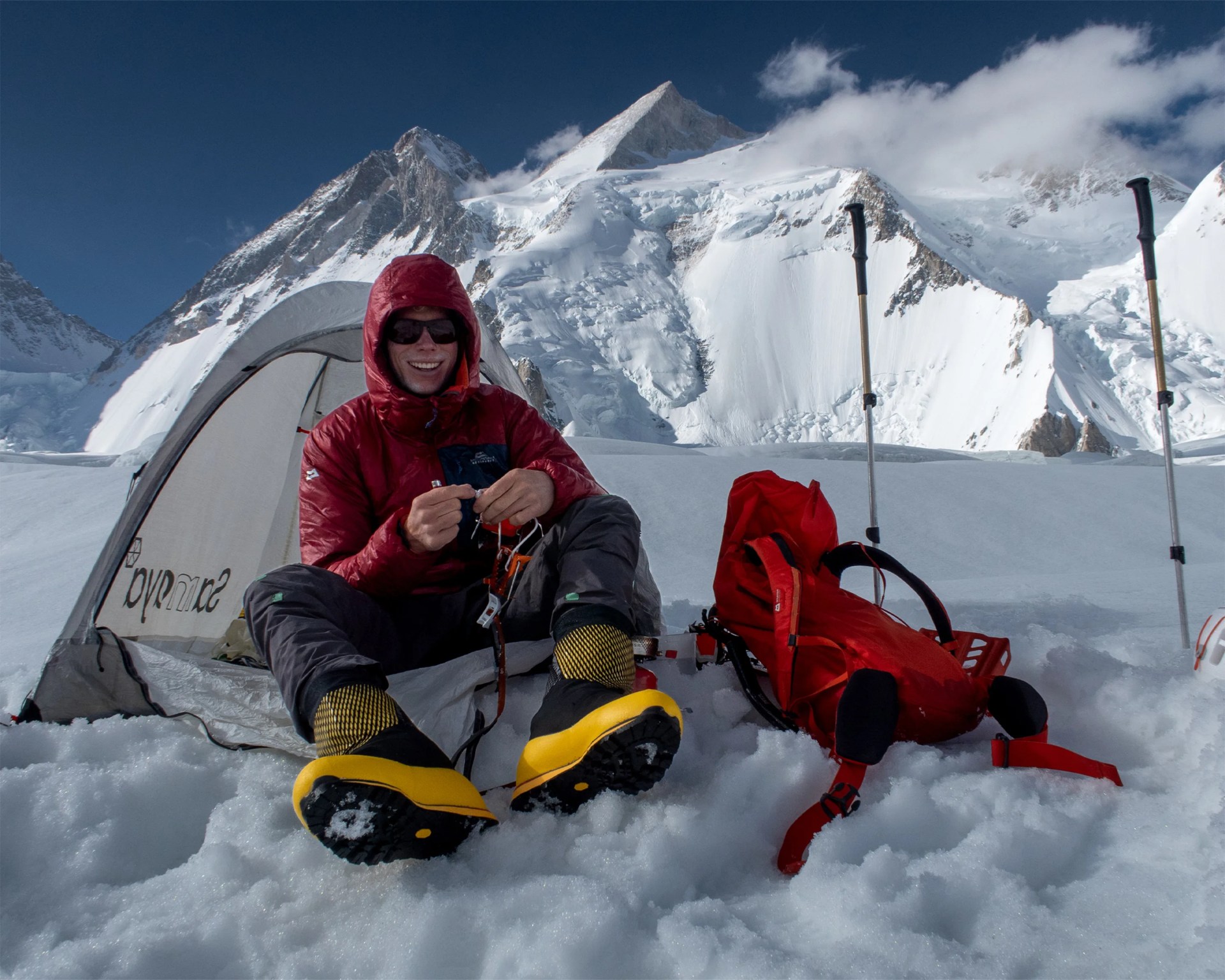 Person in a red jacket and yellow mountaineering boots sitting on snow outside a tent with a red backpack and trekking poles nearby.
