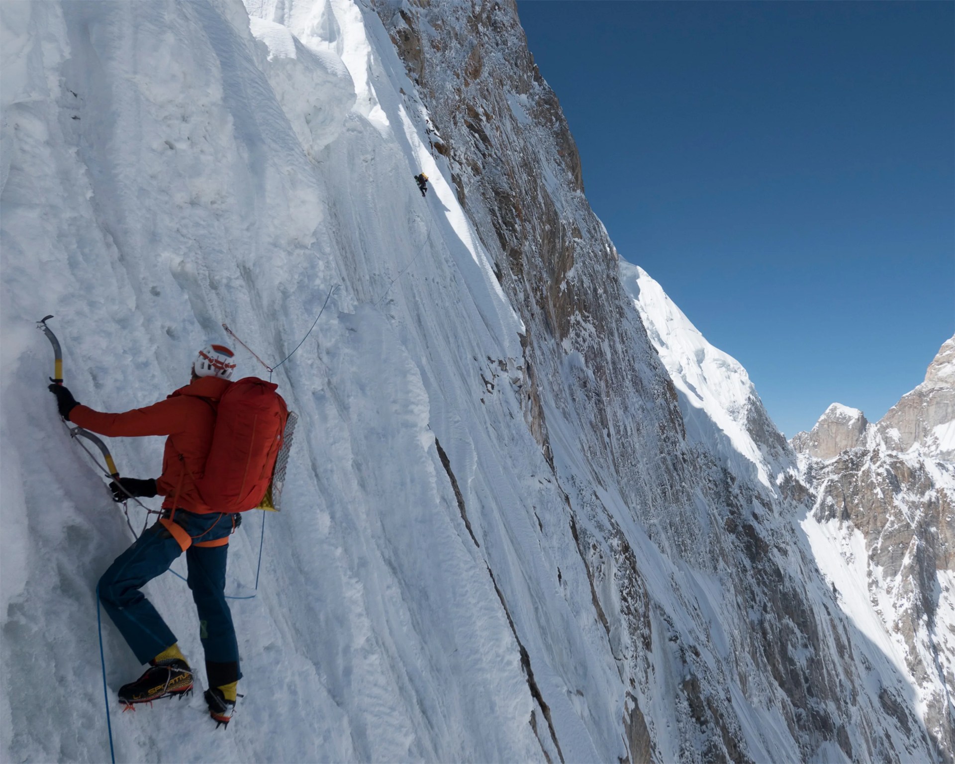 Climber in red jacket and helmet ascending steep icy mountain slope with ice axes and crampons.