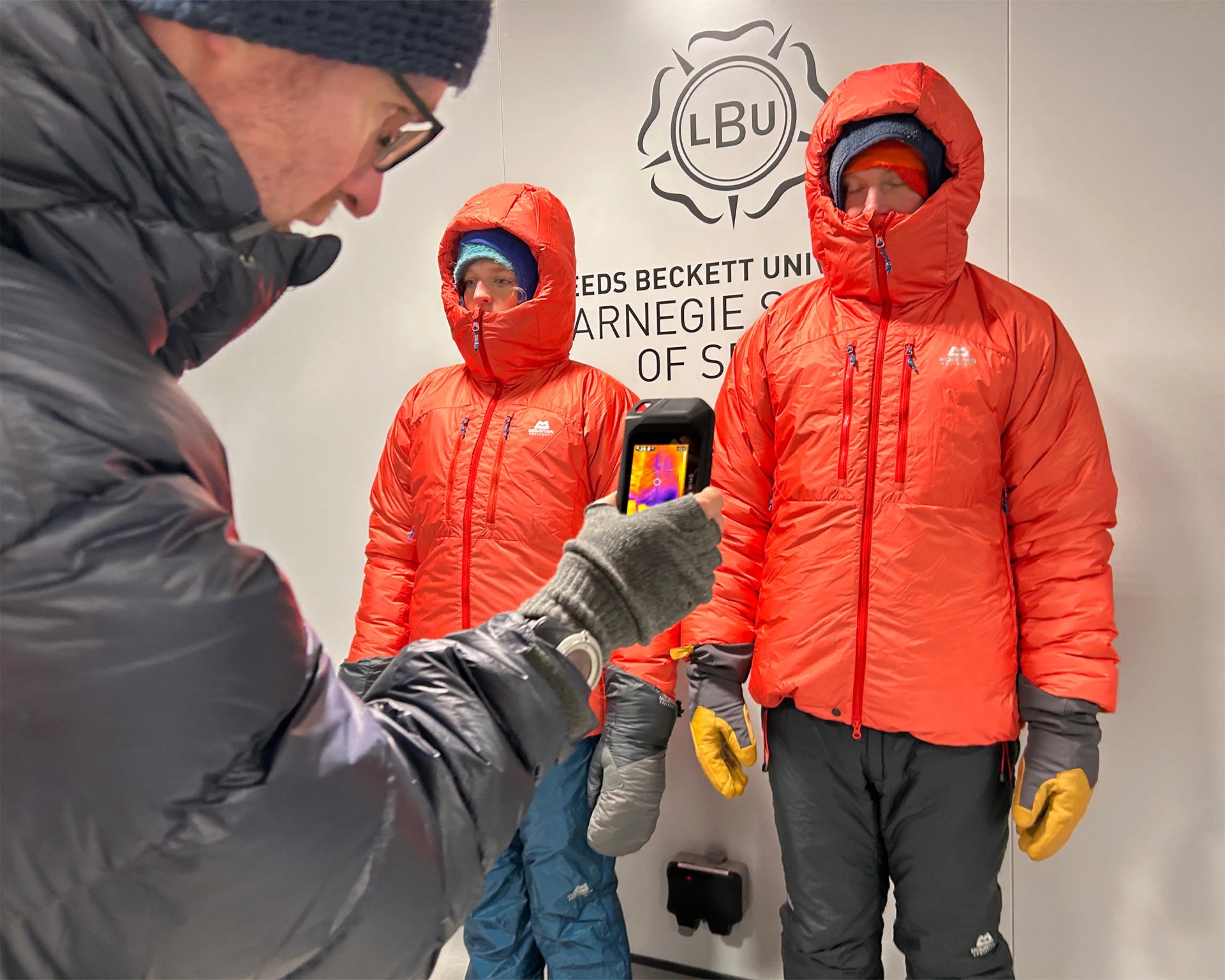 Person in black jacket and gloves holding a thermal camera pointed at two people in orange insulated jackets and gloves standing indoors.