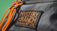 Close-up of a black fabric patch with orange "Mystery Ranch Bozeman MT USA" text on a gray bag with orange cords.