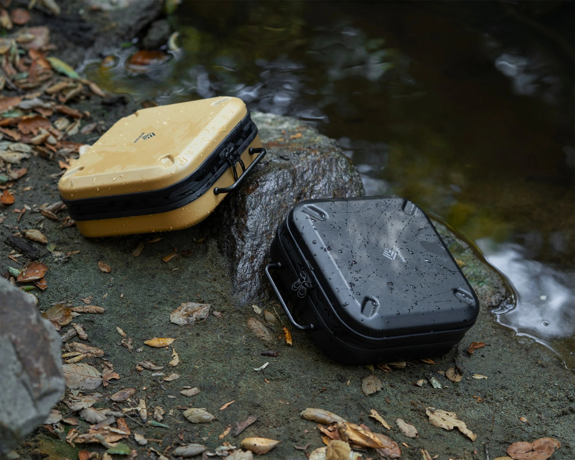 Two waterproof hard cases, one tan and one black, with handles and water droplets on them, placed on a rocky surface near water.