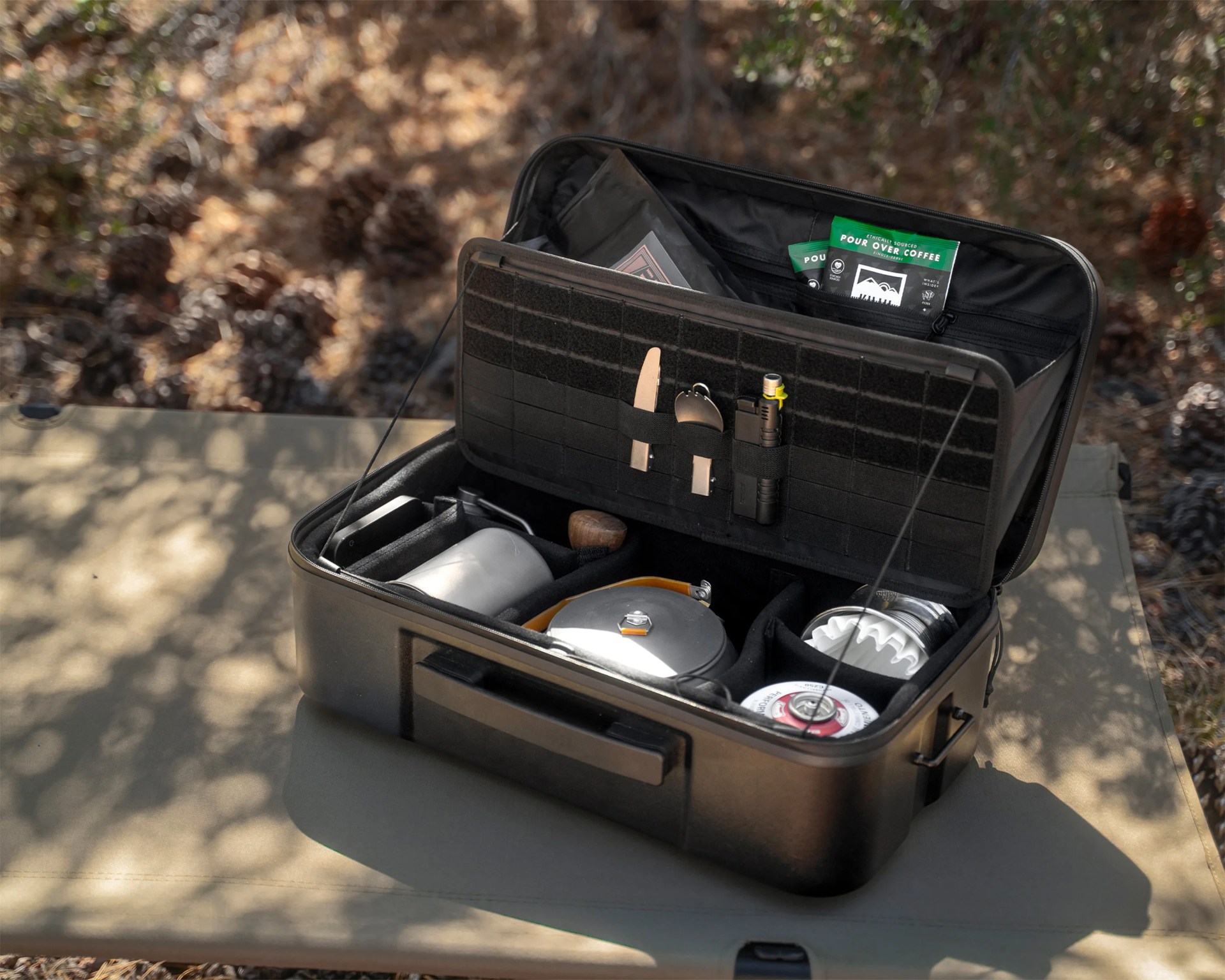 Black portable coffee brewing kit with kettle, grinder, gas canister, and utensils inside an open case on a camping cot.