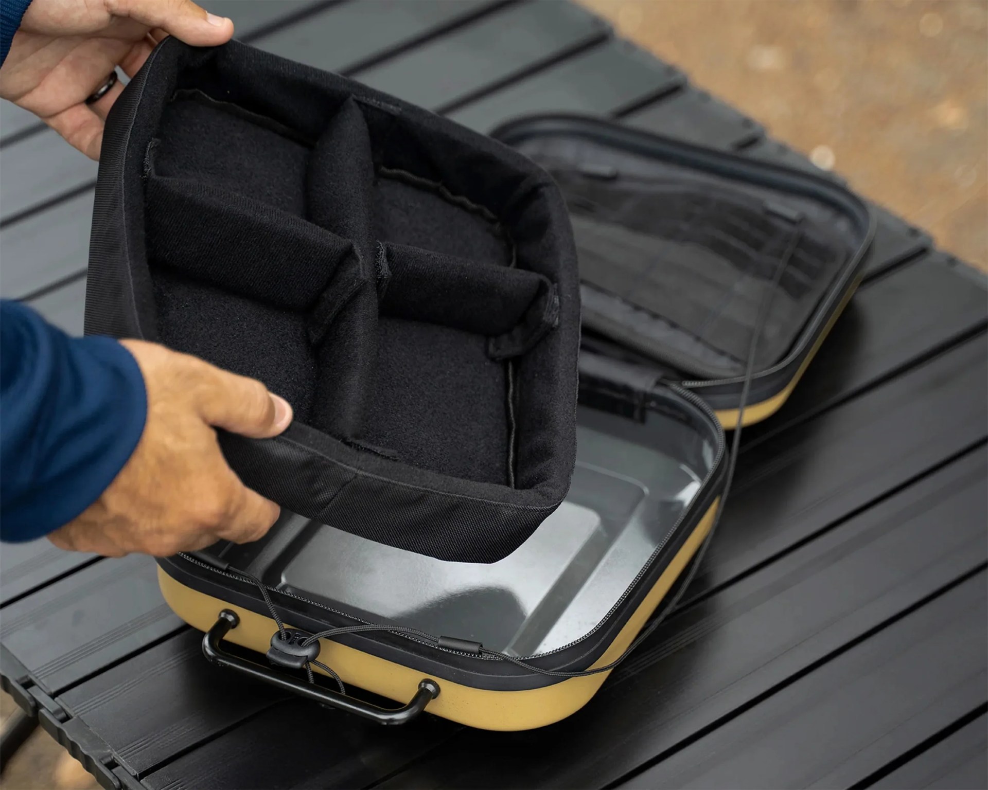 Hands holding a black padded divider above an open yellow and black hard-shell case on a black table.