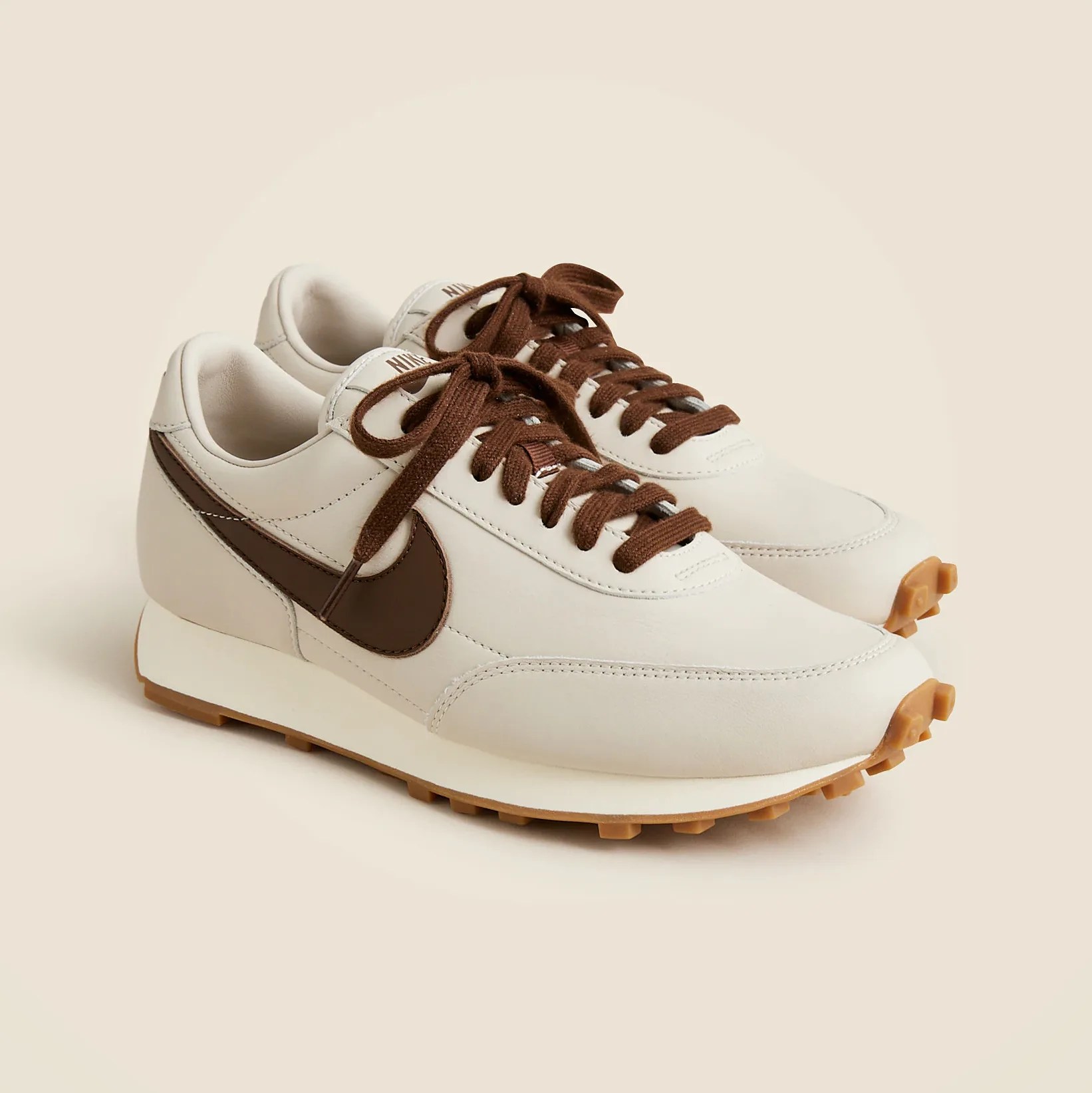 Pair of off-white Nike sneakers with brown swoosh logo, brown laces, and gum rubber soles on a beige background.