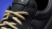 Close-up of black leather sneaker with beige woven laces against a blue background.