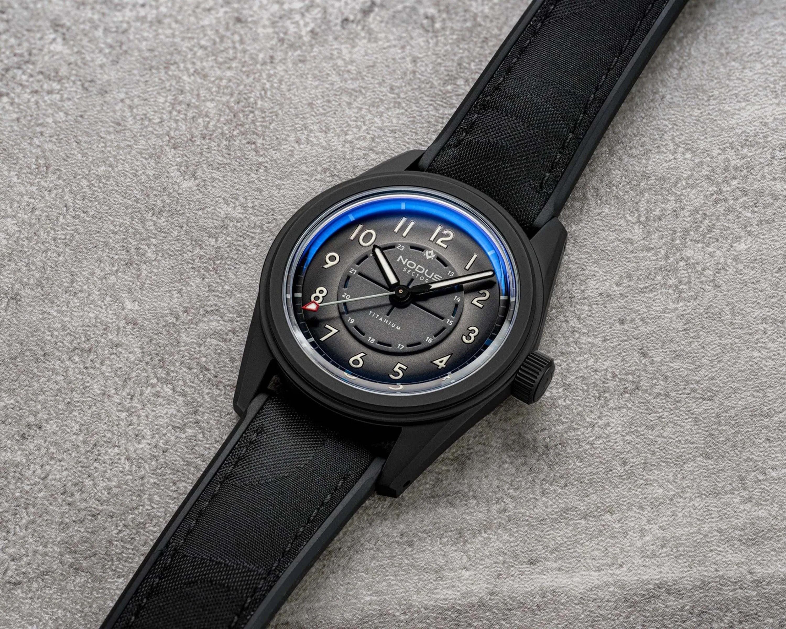 Black wristwatch with a gradient gray dial, white numerals, and a black fabric strap on a gray textured surface.