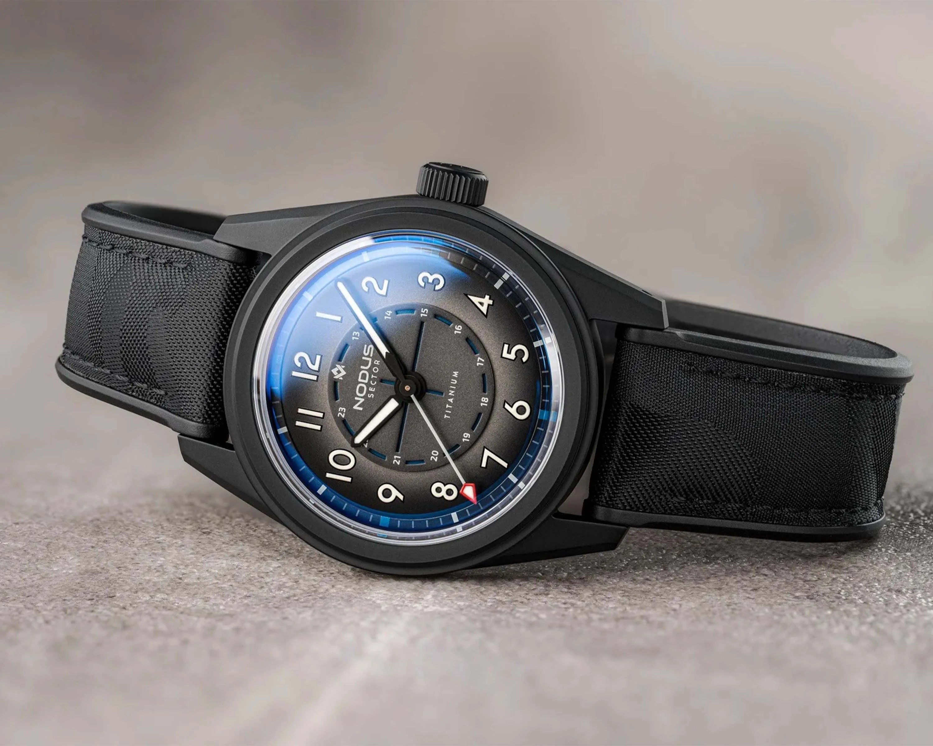 Black Nodus Sector titanium wristwatch with white numerals and black fabric strap on a gray surface.