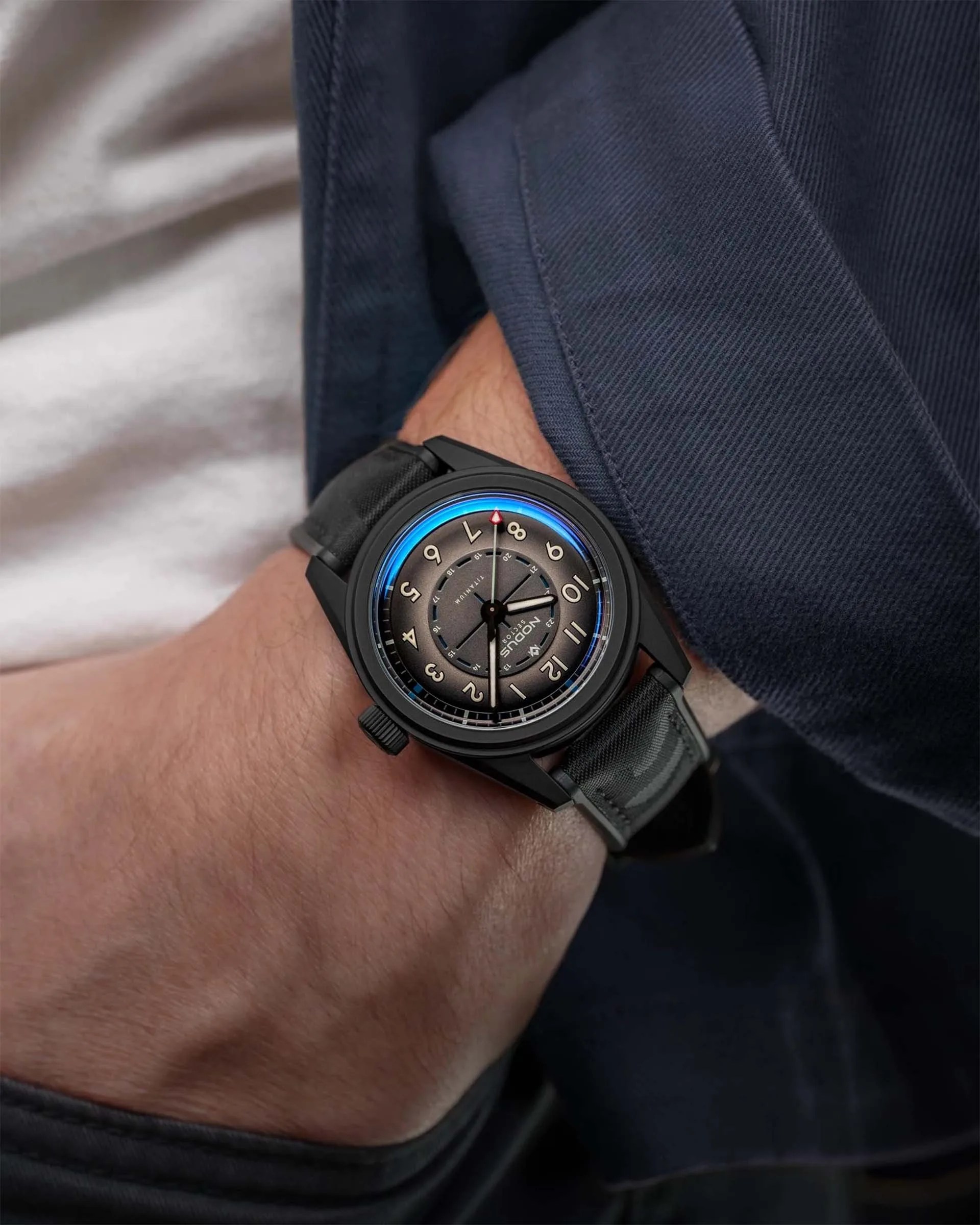 Black wristwatch with a gray dial, Arabic numerals, and a black fabric strap worn on a wrist.