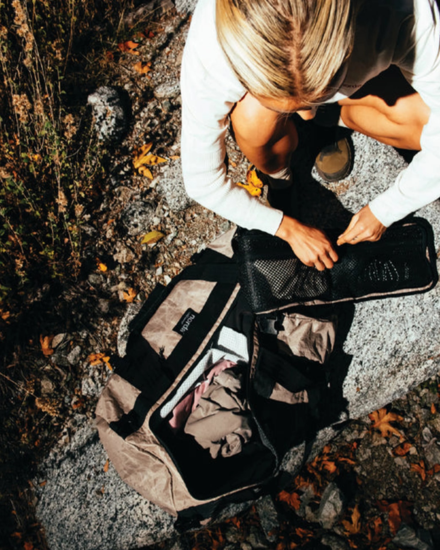 Person with blonde hair wearing a white long-sleeve shirt packing clothes into a tan and black travel bag on rocky ground.