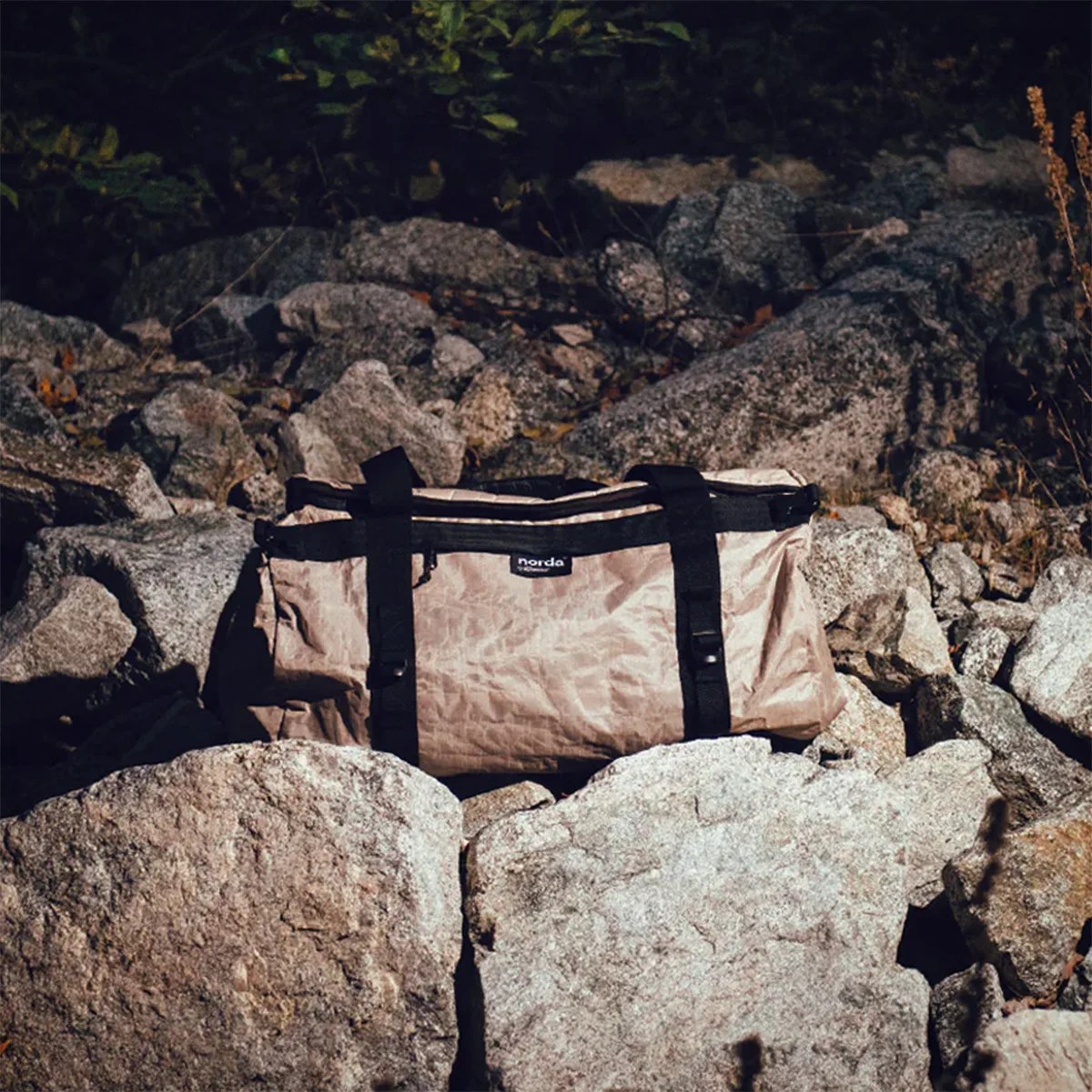 Beige duffel bag with black straps and zipper resting on gray rocks outdoors.