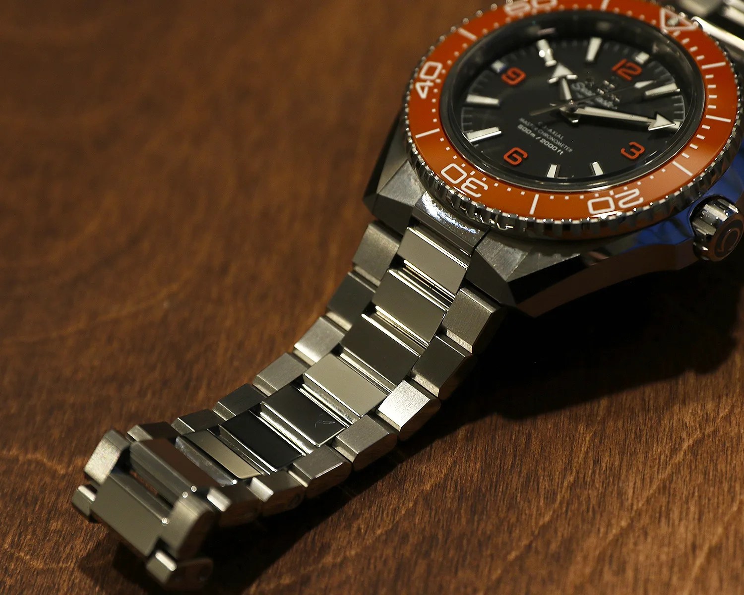 Close-up of a stainless steel watch bracelet attached to a watch with an orange bezel and black dial on a wooden surface.