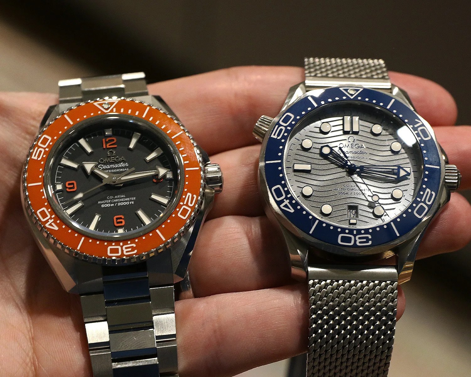 Two Omega Seamaster wristwatches held in a hand, one with an orange bezel and black dial, the other with a blue bezel and silver wave-patterned dial.
