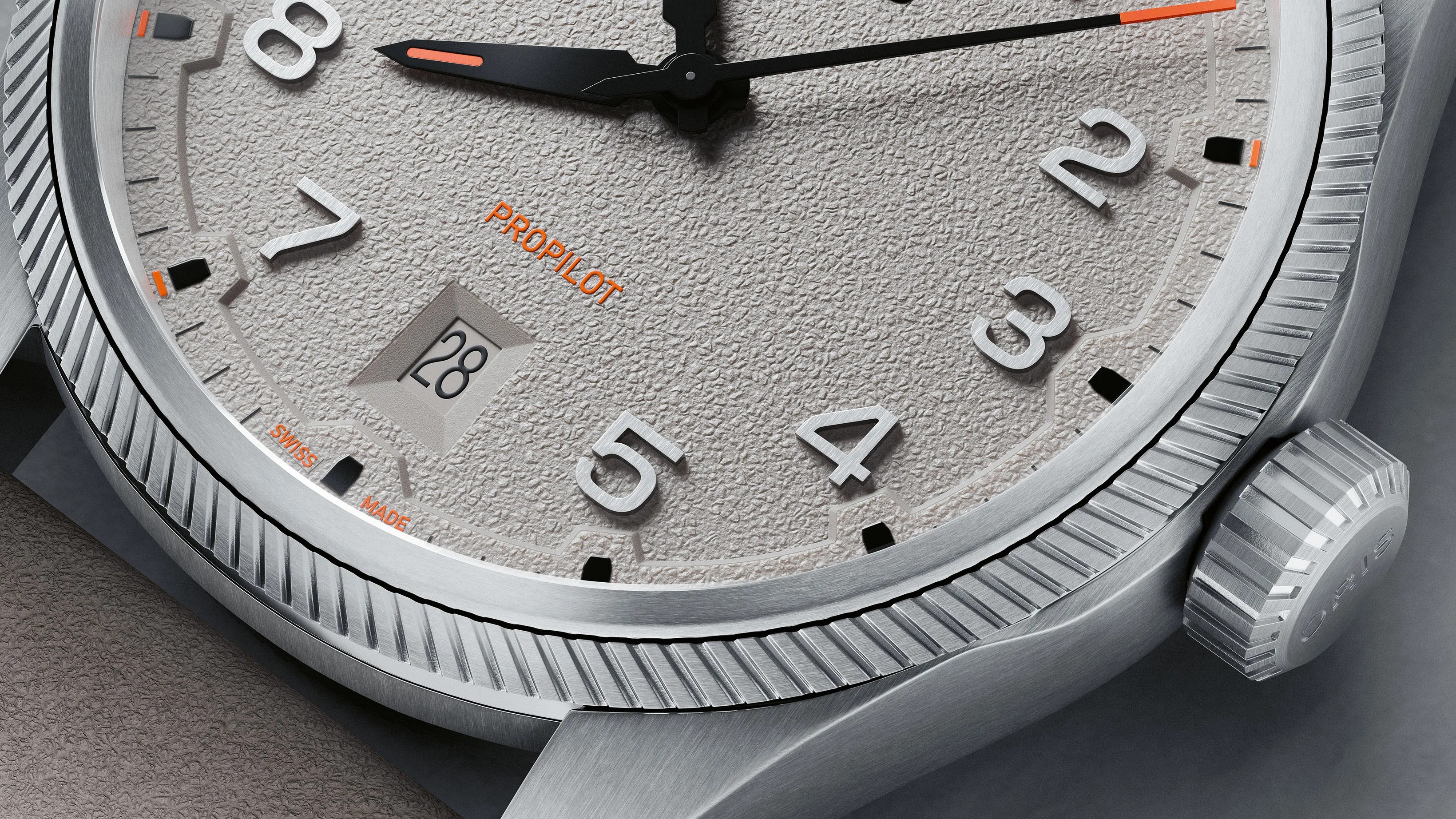 Close-up of a textured light gray watch dial with silver numerals, black and orange hands, date window at 6 o'clock, and a fluted silver bezel.