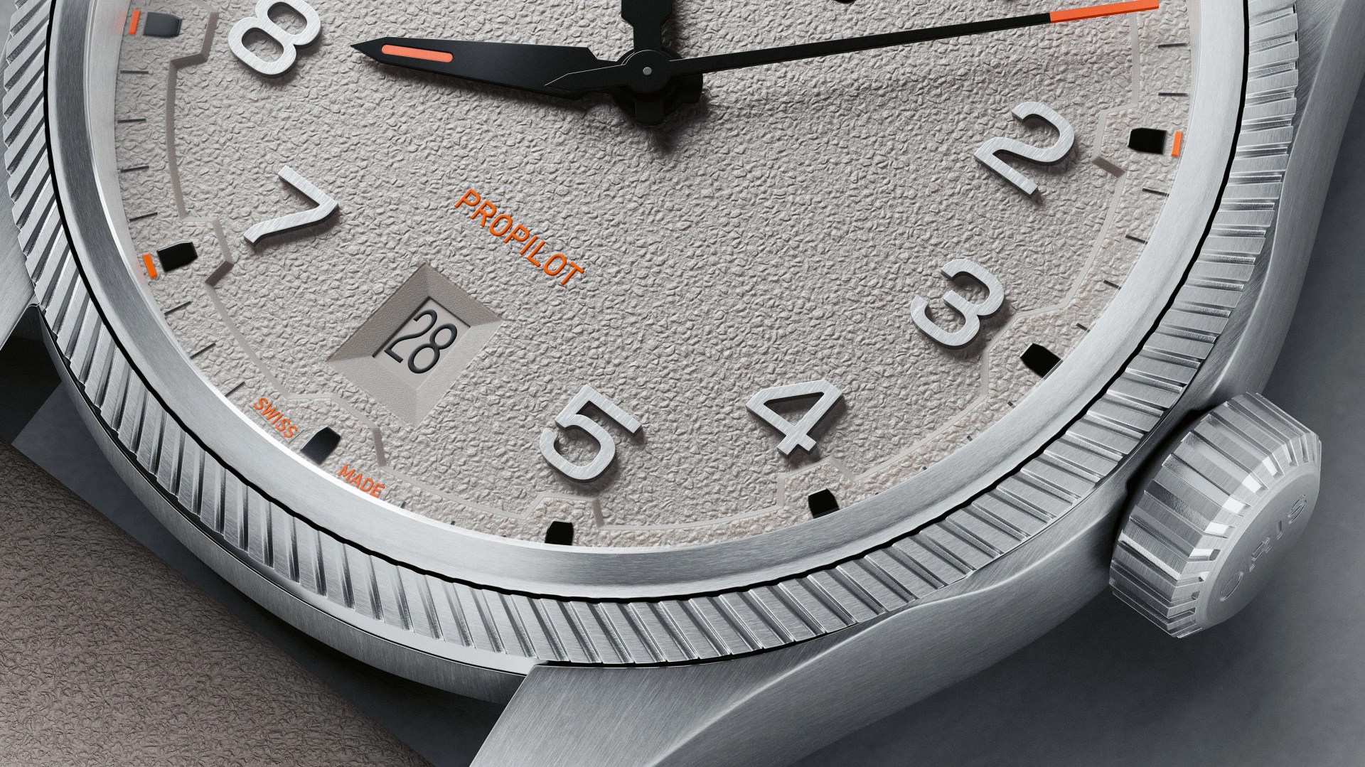 Close-up of a textured light gray watch dial with silver numerals, black and orange hands, date window at 6 o'clock, and a fluted silver bezel.