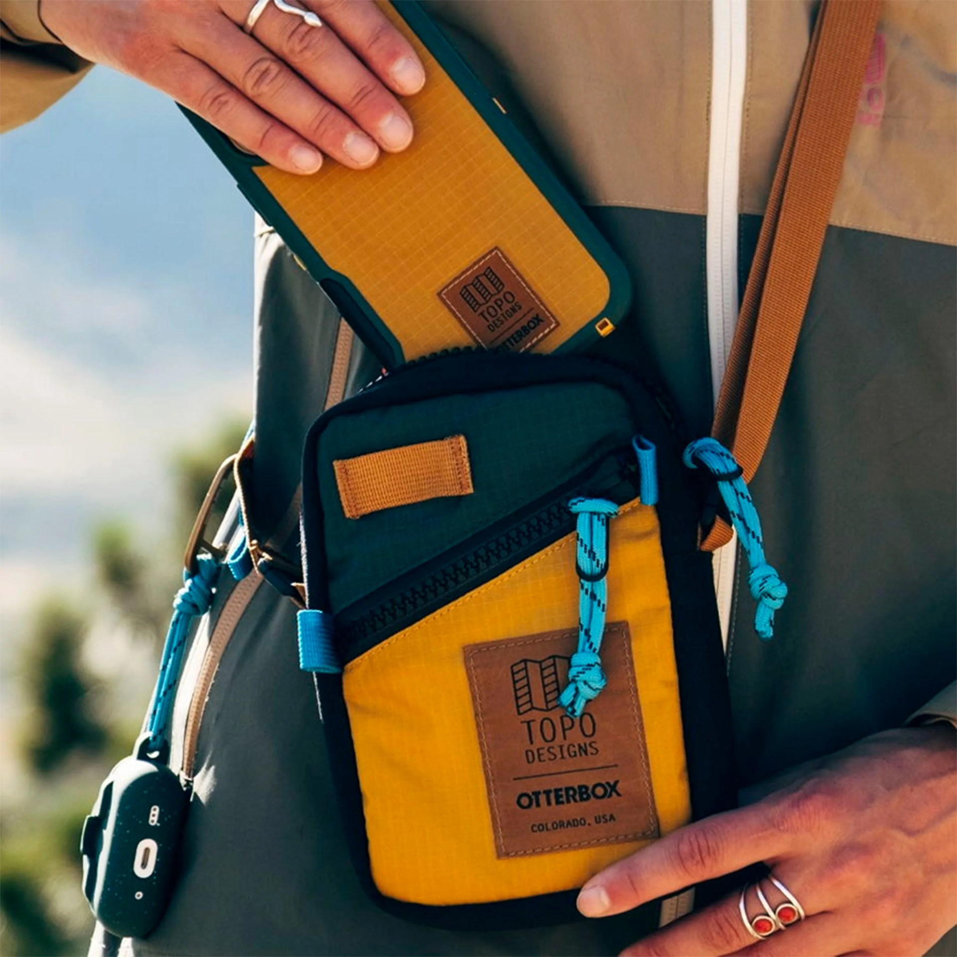 Yellow and dark green Topo Designs OtterBox crossbody bag with blue zipper pulls worn by a person.
