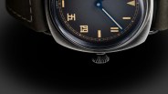 Close-up of a wristwatch with a black dial, beige hour markers, blue hands, and a brushed metal case on a dark background.