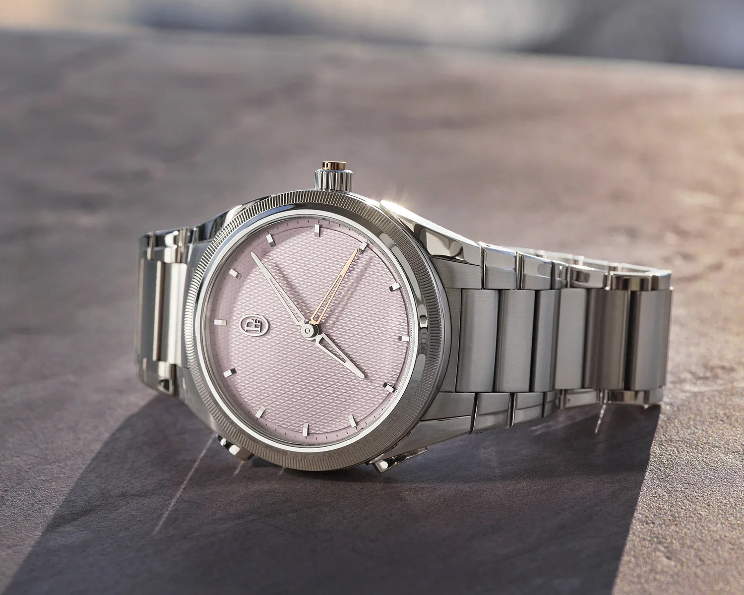 Silver metal wristwatch with a textured light pink dial and simple hour markers on a stone surface.