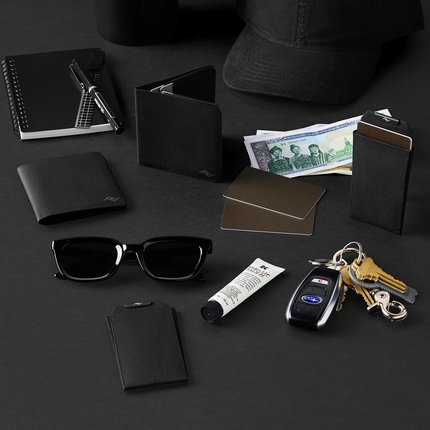 Black sunglasses, wallets, cardholders, a pen, a notebook, a tube of cream, a car key with keys, and a black cap on a black surface.