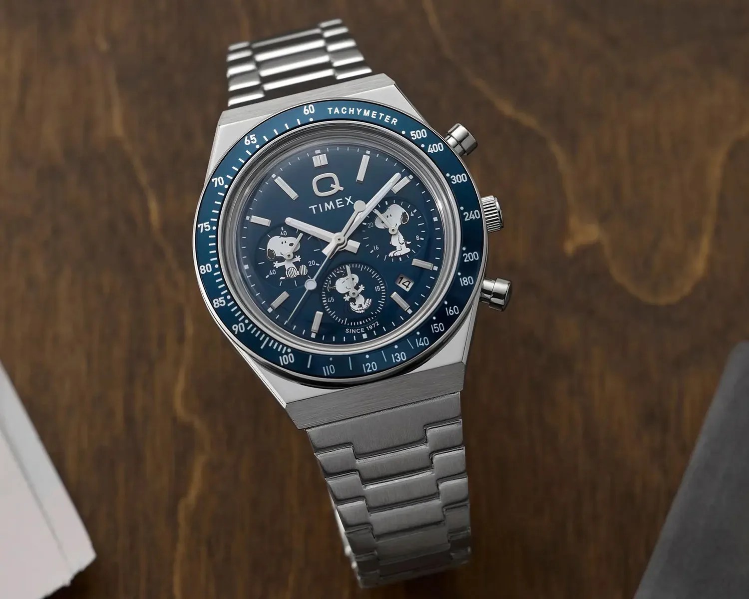 Silver stainless steel Timex Q wristwatch with blue tachymeter bezel and Snoopy character illustrations on the dial.