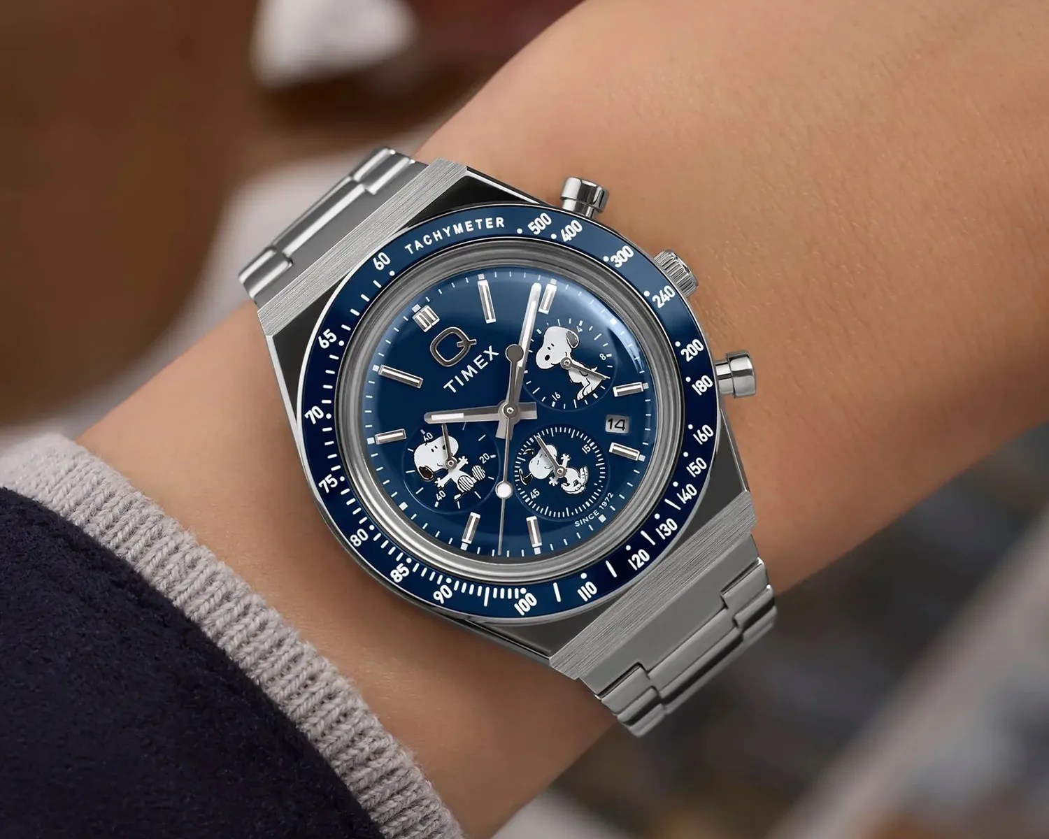 Silver metal Timex wristwatch with blue tachymeter bezel and Snoopy character on the blue dial worn on a wrist.