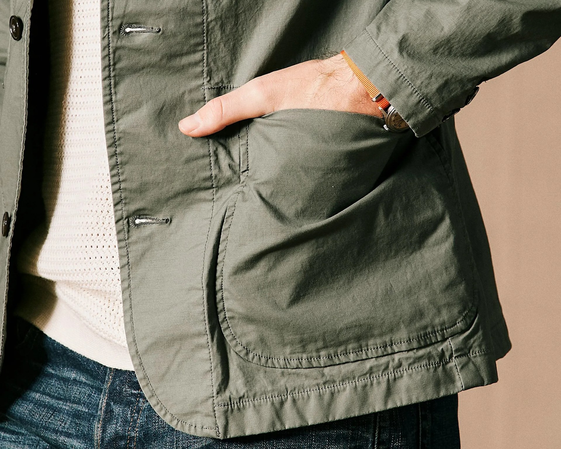 Hand in the large front pocket of an olive green jacket worn over a cream knit sweater and blue jeans.