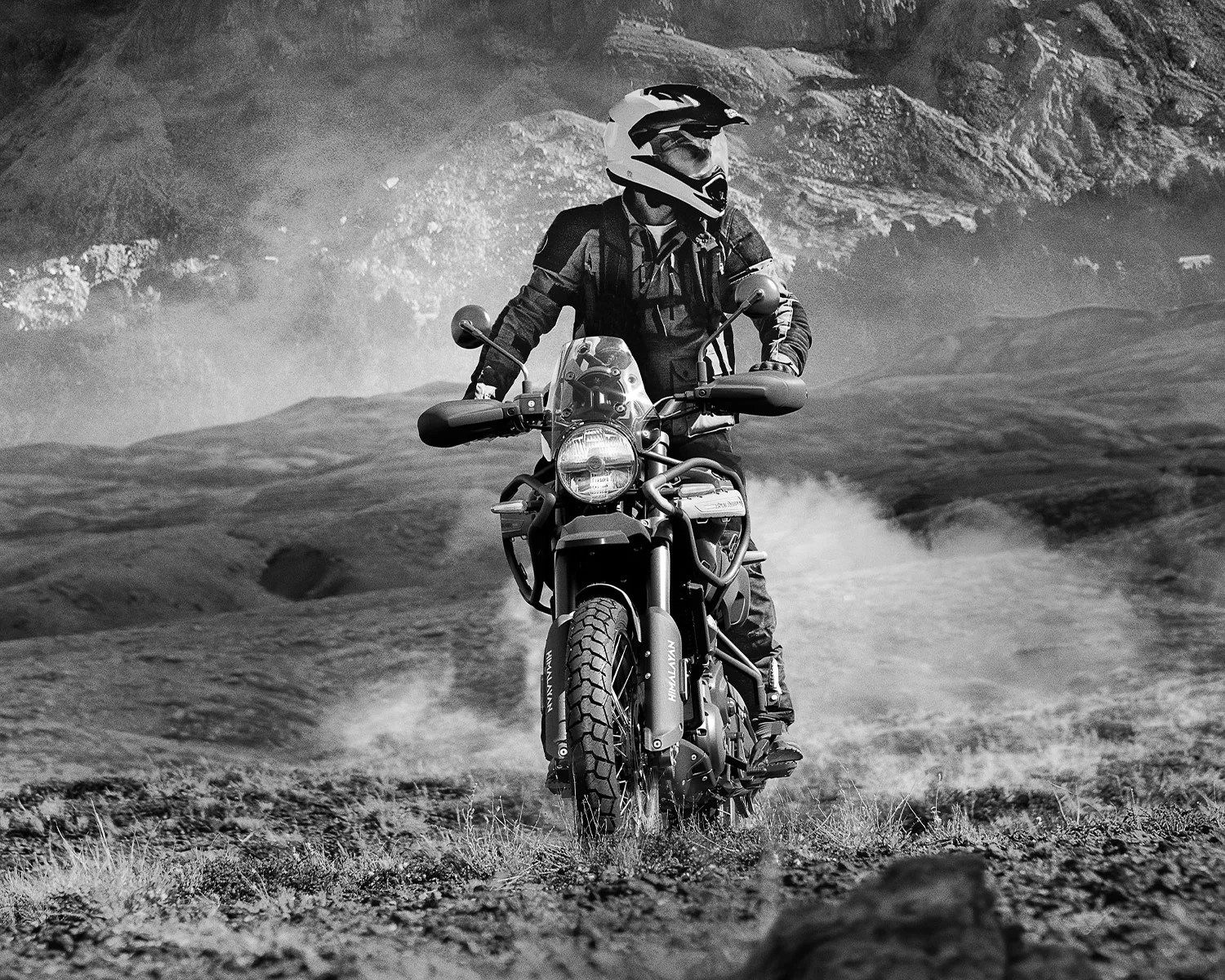 Motorcyclist wearing a helmet and gear riding a Royal Enfield Himalayan on a rugged terrain with dust clouds.