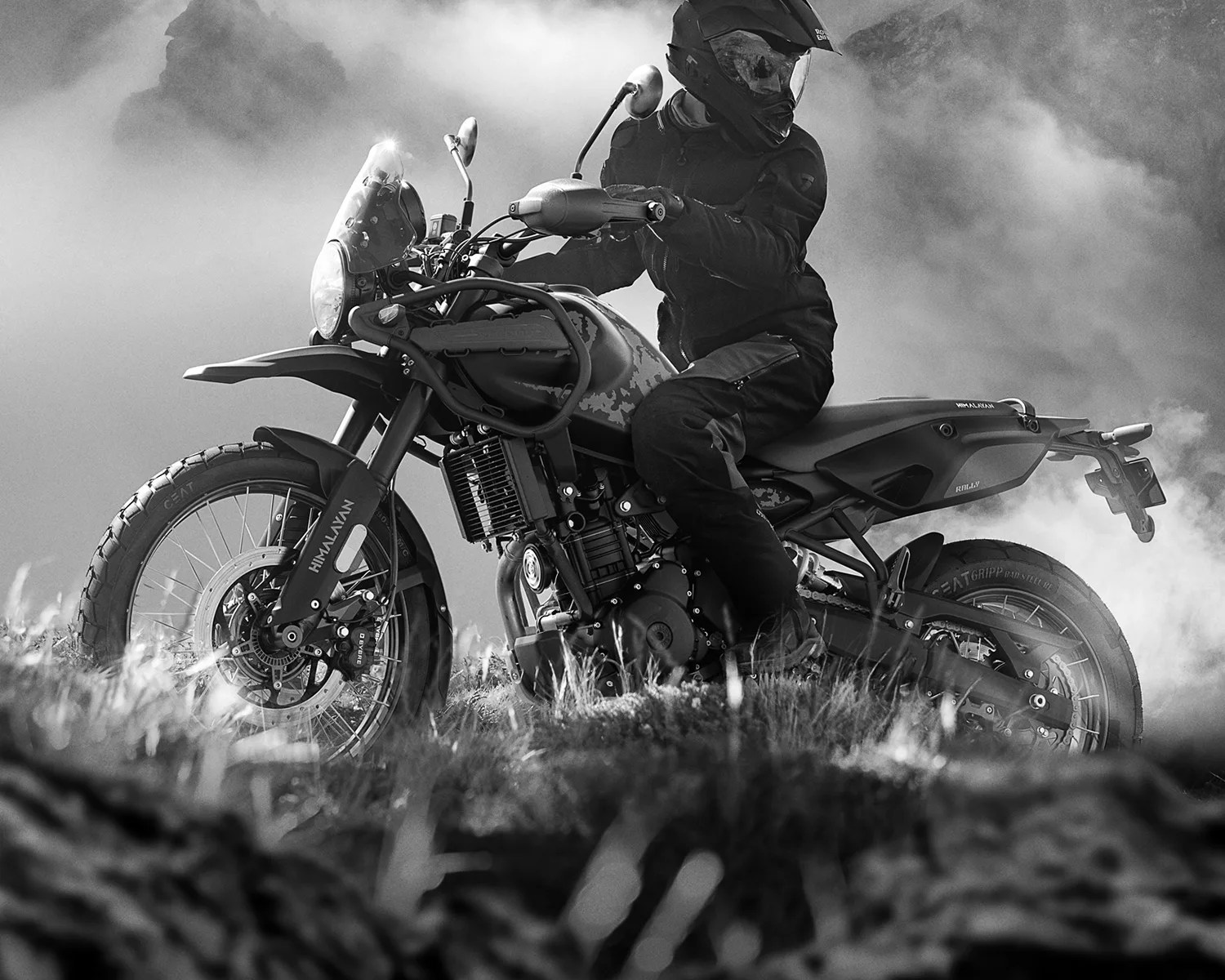 Black and white photo of a rider in full gear on a Royal Enfield Himalayan motorcycle on grassy terrain with smoke in the background.