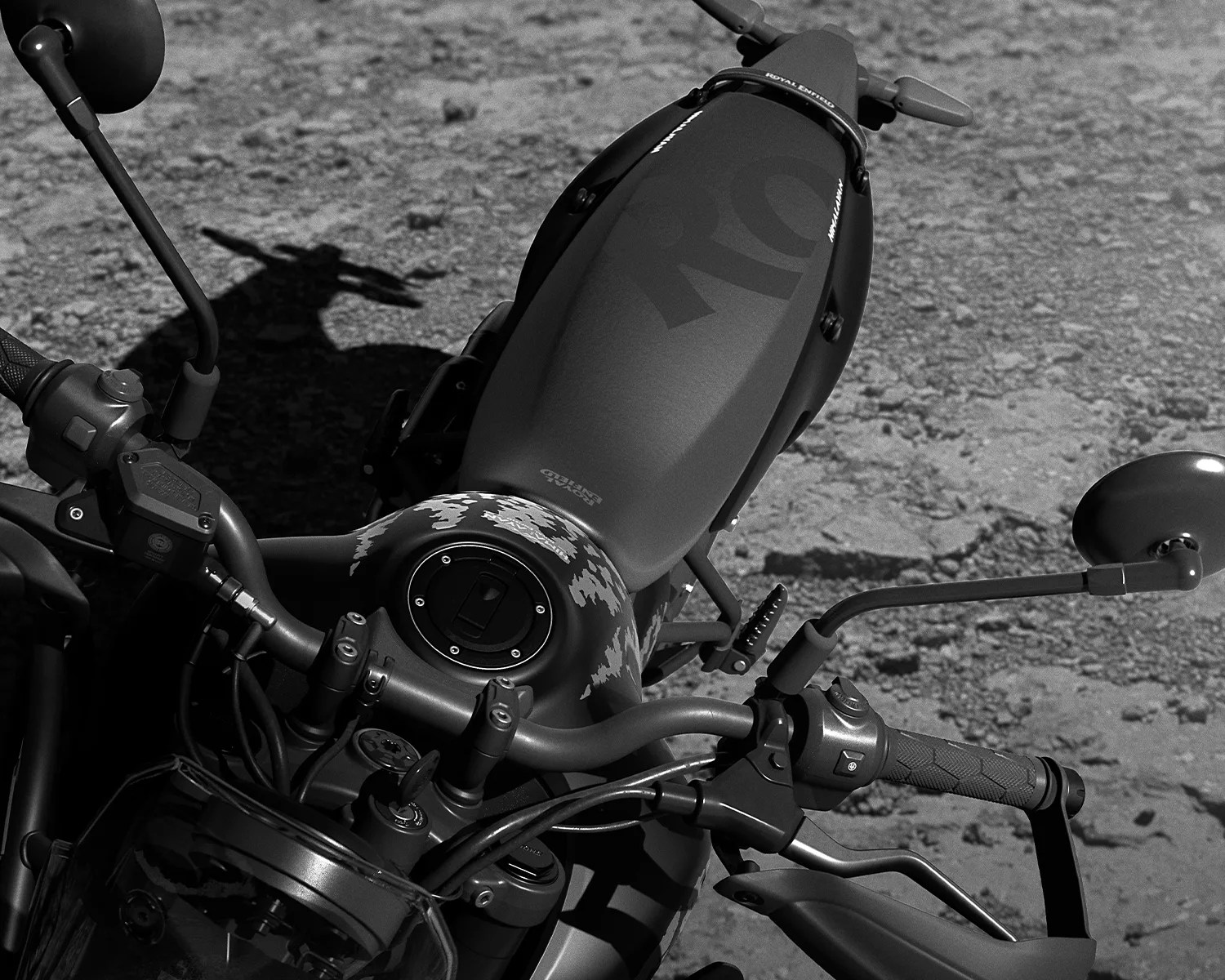 Close-up of a matte black Royal Enfield motorcycle fuel tank and handlebars on a rough asphalt surface.