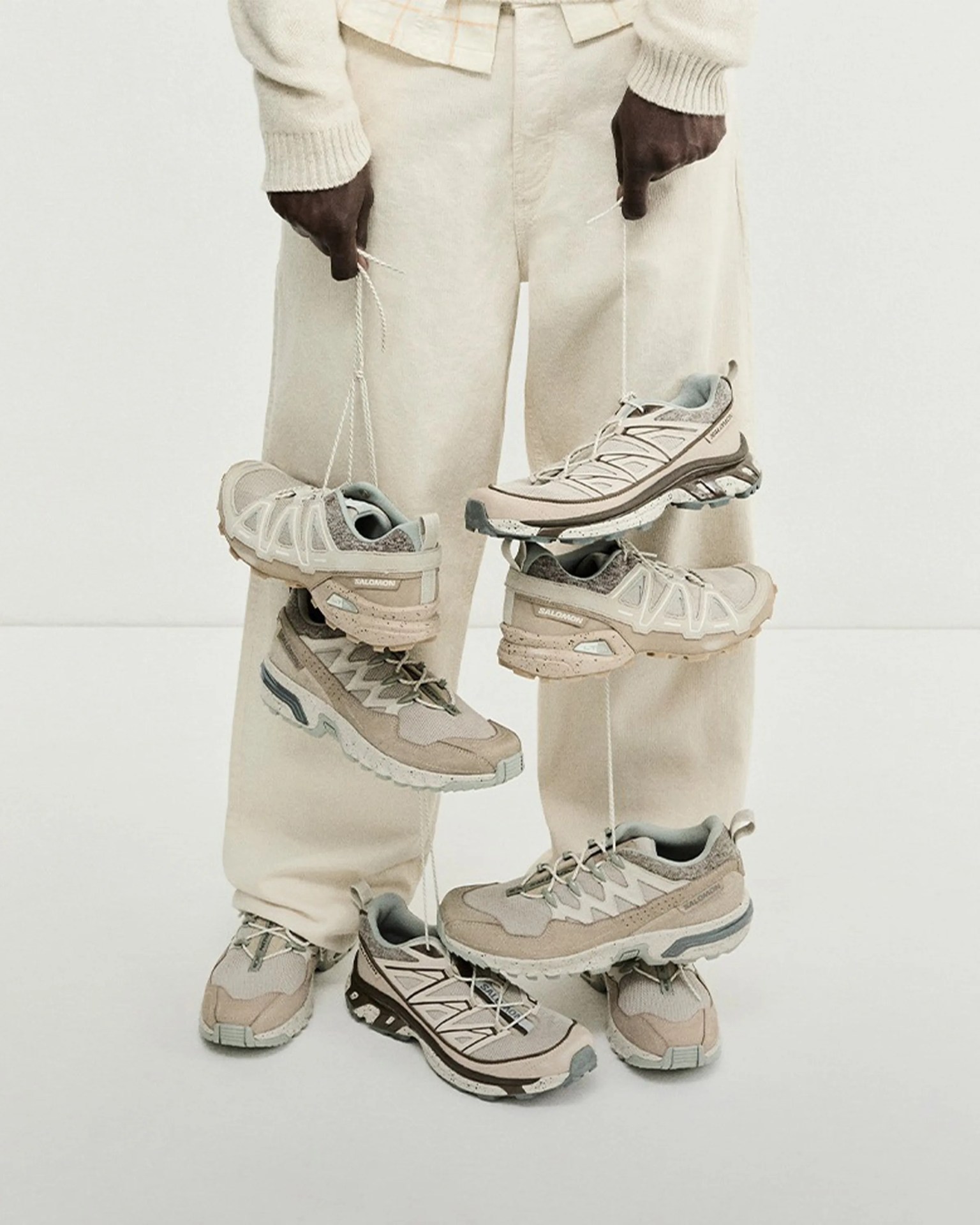 Person wearing cream pants holding multiple pairs of beige and gray Salomon trail running shoes by their laces.