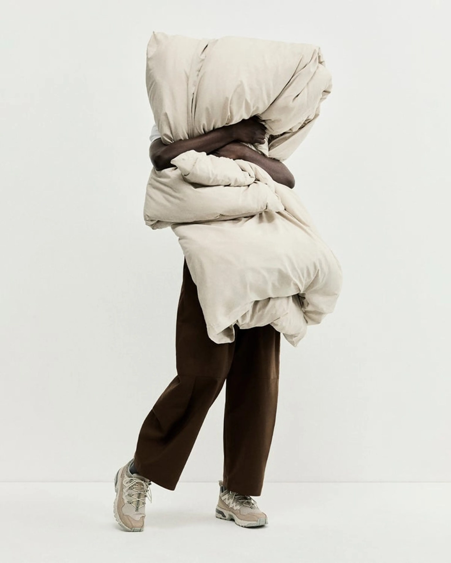 Person holding a large beige comforter, wearing brown pants and beige sneakers against a white background.